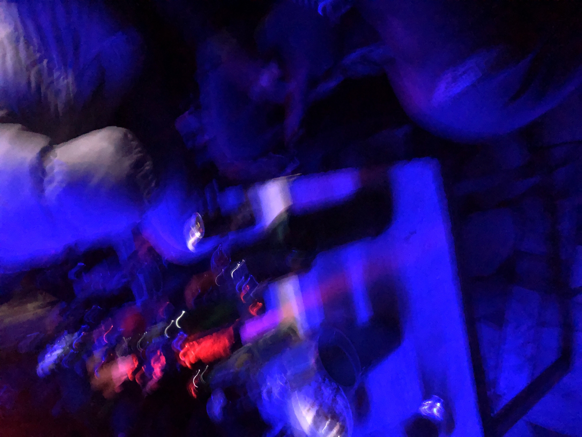 A heavily motion-blurred and low-light photograph depicts an indoor social setting, likely a bar or club, at night in the city of Unknown, Unknown. The scene is dominated by deep blue ambient lighting, with prominent streaks of vibrant red and wavy white light trails, indicative of moving light sources and camera shake. In the lower half, a light-colored table is visible, upon which several dark-colored bottles, consistent with beer or spirit containers, and at least one round, light-colored bucket or bowl are present. Two indistinct human figures are discernible within the blue and red lighting: one heavily blurred upper torso or head is in the upper right quadrant, and another figure with a visible arm reaching across is in the mid-right. The extensive motion blur renders specific actions and interactions ambiguous, but the overall environment suggests a dynamic social gathering with people and beverages.