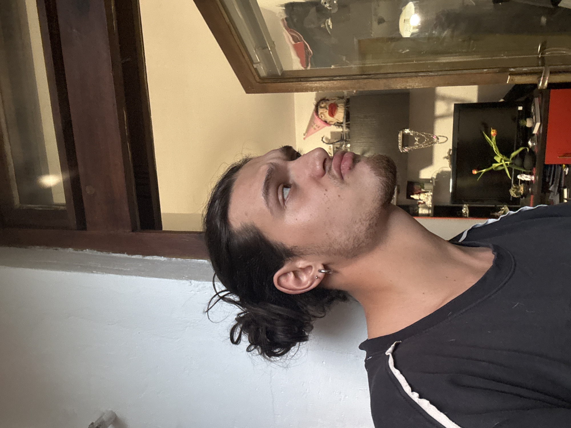 A man with long dark hair pulled back into a bun is looking upwards. He is wearing a black shirt with white stripes. The background includes a window and a display case containing various objects. The location cannot be determined from the image alone, and there is no evidence to suggest it is in Sofia, Bulgaria.
