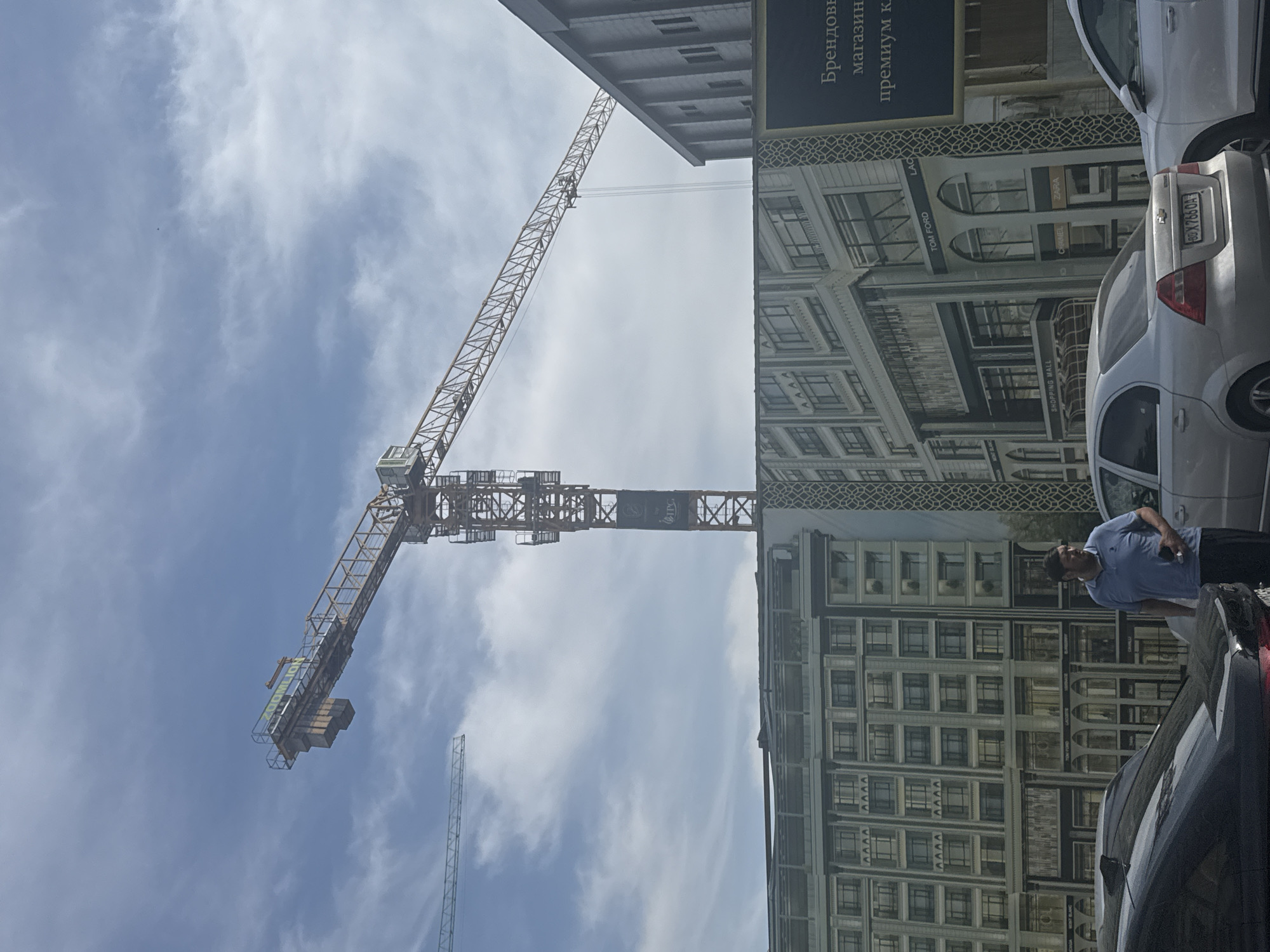 The image shows a construction scene in Tashkent, Uzbekistan. A large crane dominates the view, with its arm extending across a partly cloudy sky. A building facade is visible, draped with a promotional sign advertising premium brand stores. Cars are parked near the building. A man wearing a blue shirt is standing next to one of the cars, partially obscuring the view of the building's facade. The license plate of the car is BD X7660A. The building's facade displays the names of various luxury brands, including TOM FORD, ZARA, and CHANEL, as well as the phrase "SHOPPING MALL" and Cyrillic text reading "Брендовы магазин премиум к".