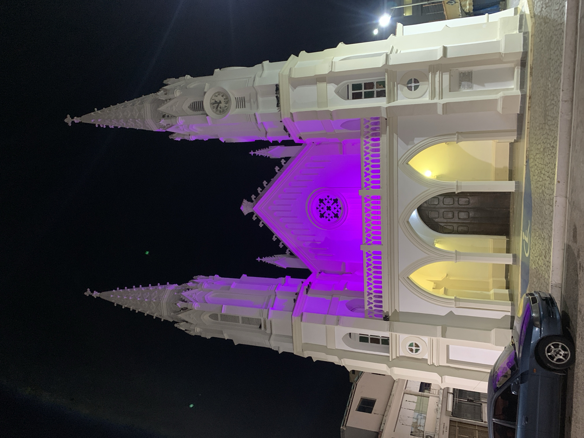 The image shows the Iglesia de San Juan Evangelista in Juan Griego, Venezuela, at night. The church is white with two tall spires and is illuminated with purple lighting on the facade and spires. A blue car is parked on the right side of the image, next to the church's entrance area. The sky is dark. Other buildings are visible adjacent to the church.