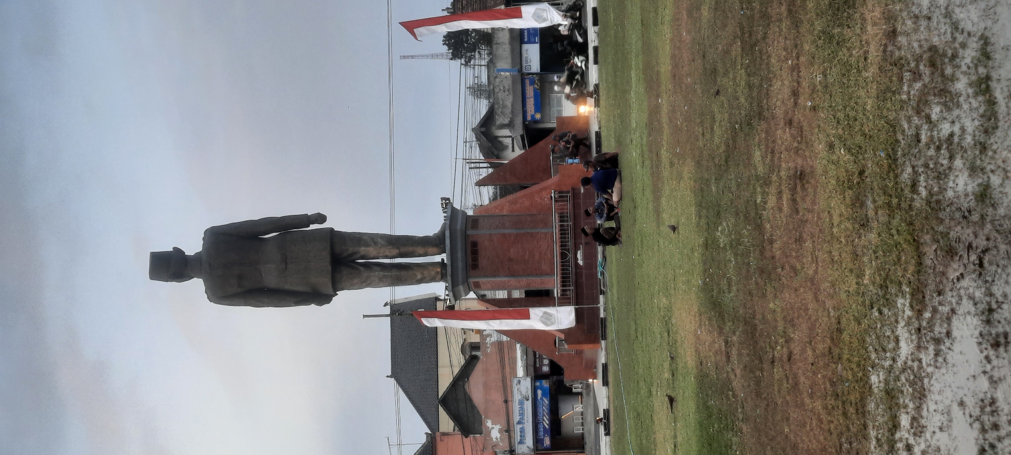 The image depicts a large statue of a man in a suit and cap, facing away from the camera. The statue stands on a brick pedestal in what appears to be a public square or park area. In the foreground, a grassy field with patches of dry grass and some puddles of water is visible. Several people are sitting and conversing on the grass near the base of the statue. In the background, there are buildings, some with signs, and a few motorcycles parked or being ridden. Two Indonesian flags (red and white) are prominently displayed on poles near the statue. The sky is overcast. The location is identified as Kanigoro, Indonesia.