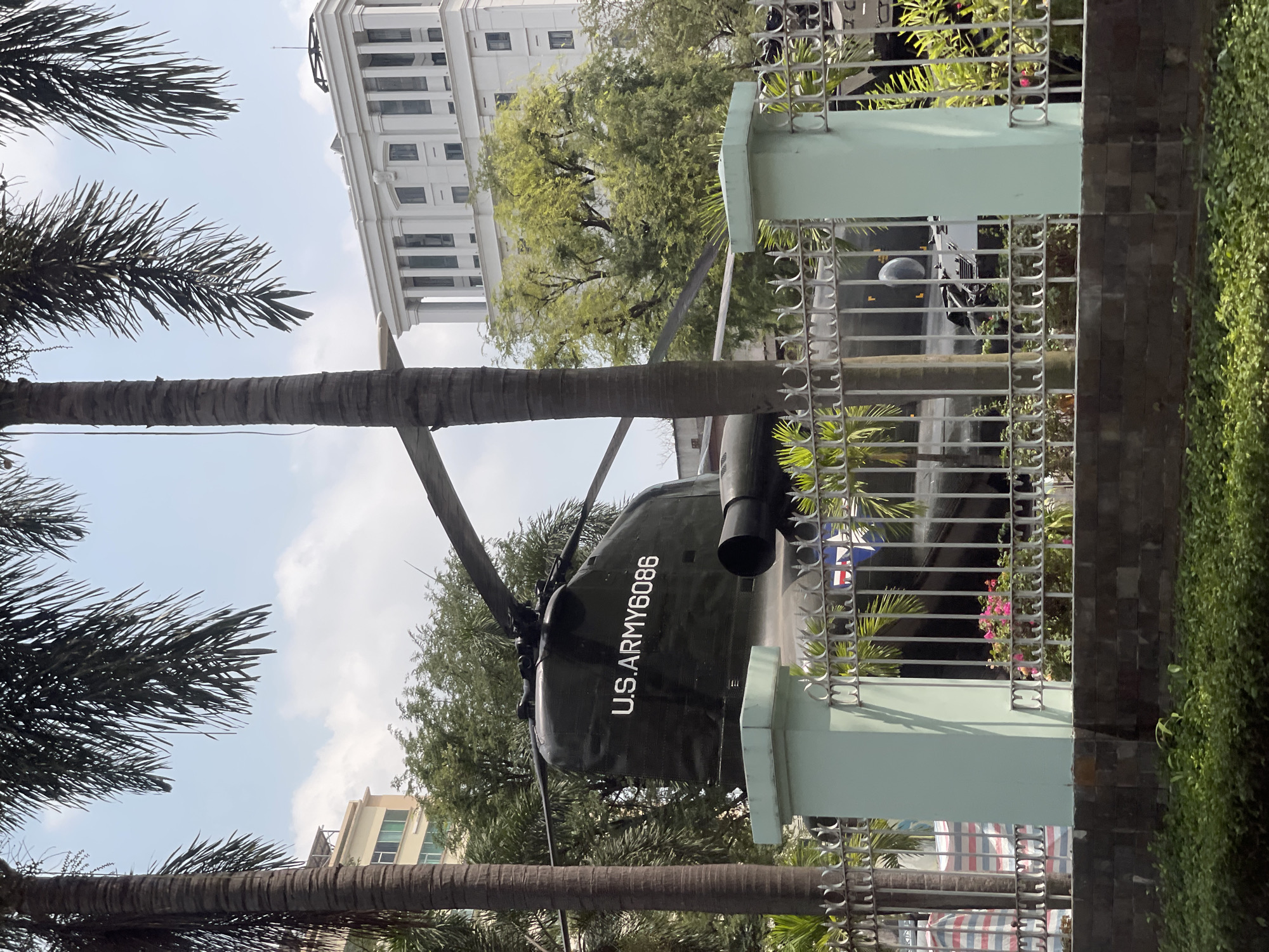 Captured in the city of Thủ Đức, Vietnam, the image presents a partially obscured view of a U.S. Army helicopter, marked with "U.S. ARMY 6086", positioned behind a metal fence with light-green pillars. The backdrop includes a multi-storied white building with classical columns. A palm tree trunk and foliage are visible in the foreground, contrasting with the urban environment. The scene is taken during daylight.
