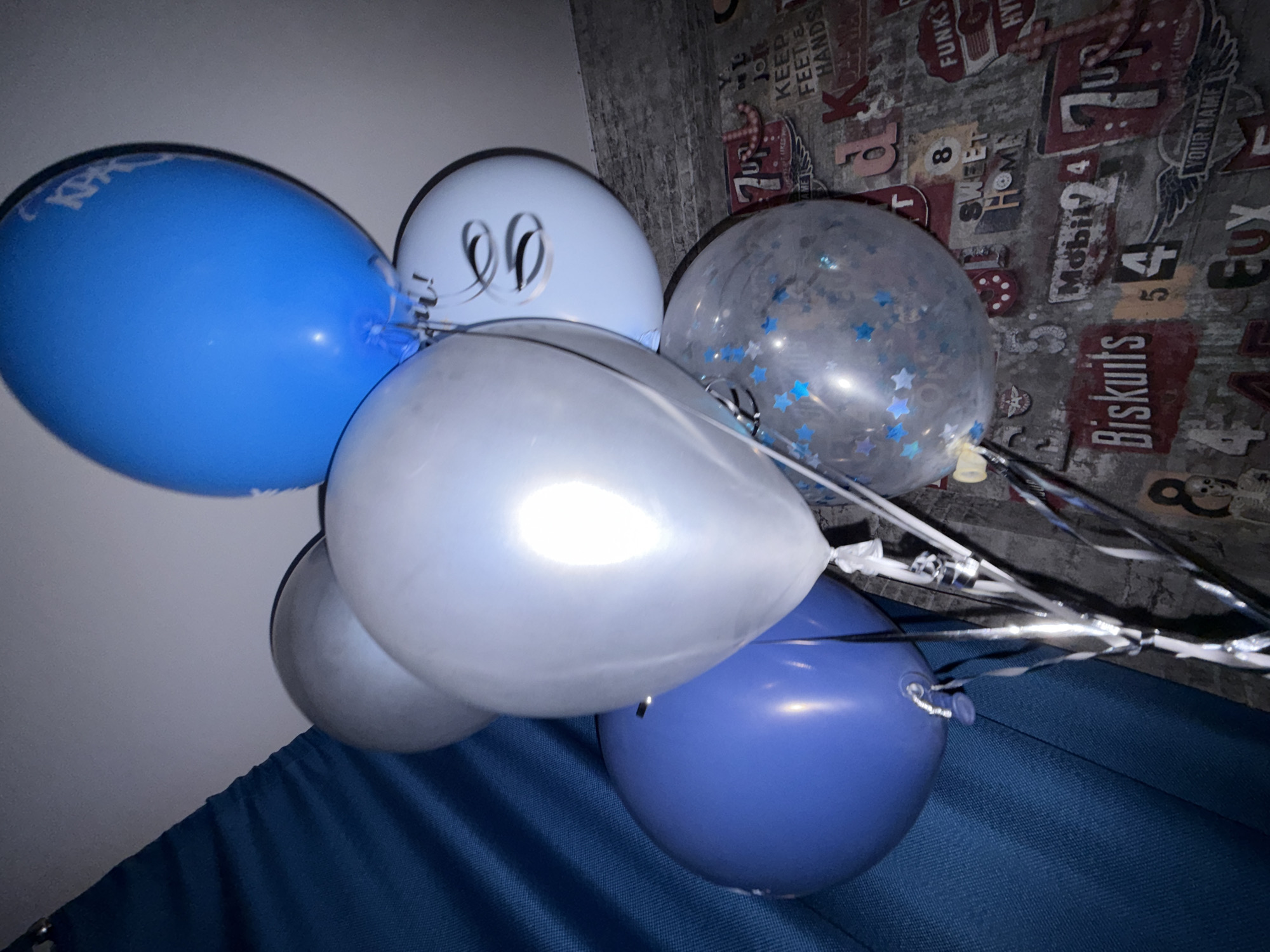 This image displays a cluster of balloons in various shades of blue and white, arranged in a close-up shot. The balloons are the primary objects, with a large bright blue balloon on the left, a lighter blue balloon with "90" written on it, a clear balloon filled with blue star confetti, and two metallic silver balloons. Some balloons have reflective ribbons attached. The background is a textured wall with a pattern that resembles vintage signage and text, including phrases like "KEEP FEET HANDS," "SWEET HOME," "YOUR NAME," and brand names like "FUNK'S" and "Biskuits." The lighting suggests an indoor setting, possibly a room or a party venue, with the balloons positioned in the foreground against this detailed background. The overall impression is festive, likely related to a celebration or birthday, given the balloons and celebratory text. There are no people visible in the image. The scene is static, with the balloons simply arranged together. The colors are somewhat muted due to the lighting, but the balloons themselves are vibrant.