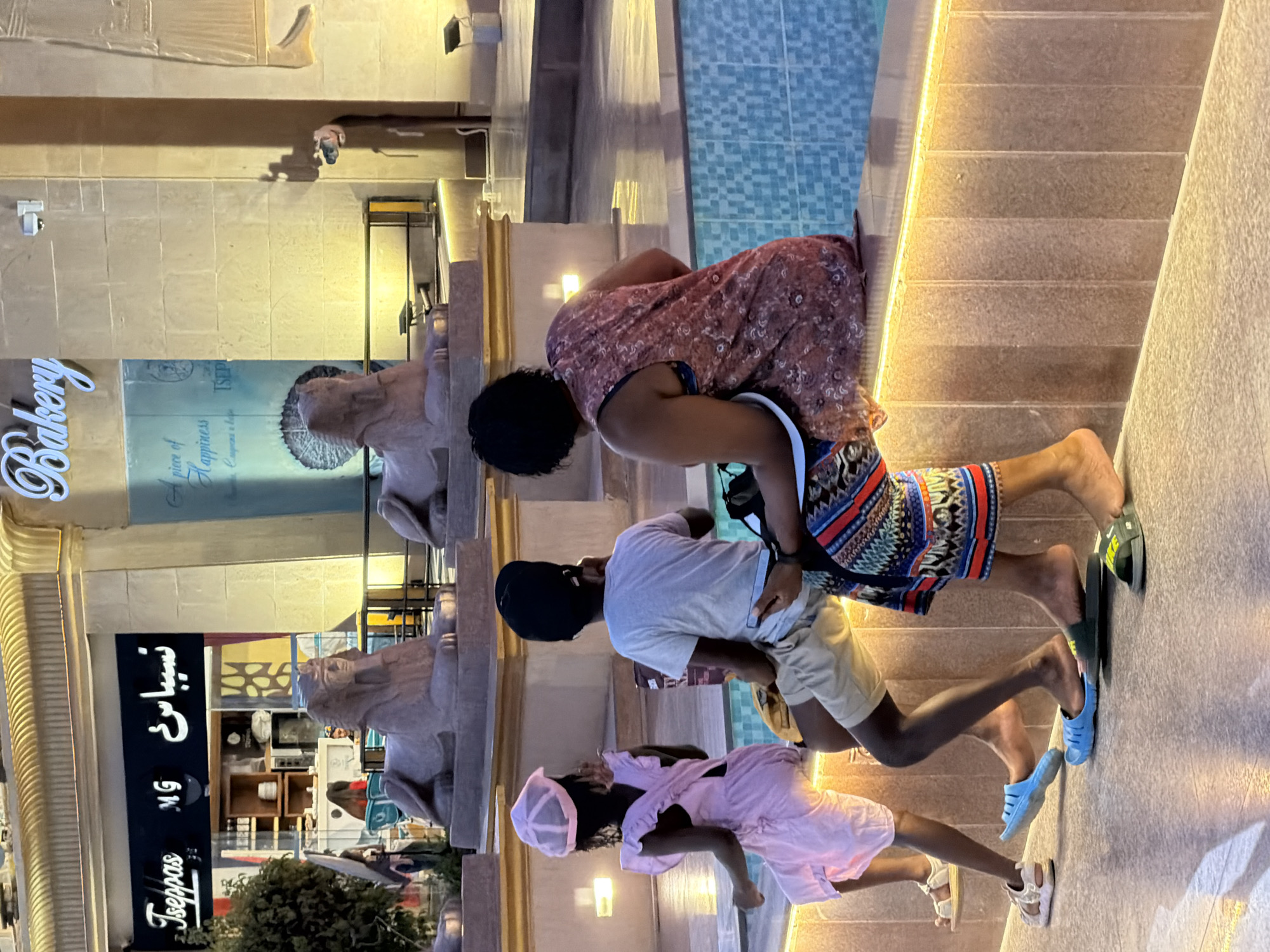 The image depicts a family of four sitting on a low wall in an outdoor public space, likely a promenade or plaza in Sharm El Sheikh, Egypt. In the foreground, a young girl wearing a pink dress and a matching hat sits on the left. To her right, a young boy in a white t-shirt and khaki shorts is seated. Next to him, a woman in a floral-patterned dress is also seated. Partially visible on the far right is another individual. The family appears to be resting, with the children looking towards the left and the adults looking away from the camera.
The background reveals architectural elements suggestive of a tourist destination. A sign with Arabic script and the English word "Tseppas" is visible on the left, accompanied by a "MG" logo. Further to the right, a sign reads "Bakery." Two stone sphinx statues, characteristic of ancient Egyptian art, are positioned prominently in the mid-ground, facing the viewers. The area also features a body of water with a mosaic tile pattern, likely a fountain or a decorative pool. The lighting suggests it is either dusk or evening, with artificial lights illuminating the scene. The overall environment appears to be a well-maintained, possibly upscale, public area within Sharm El Sheikh.