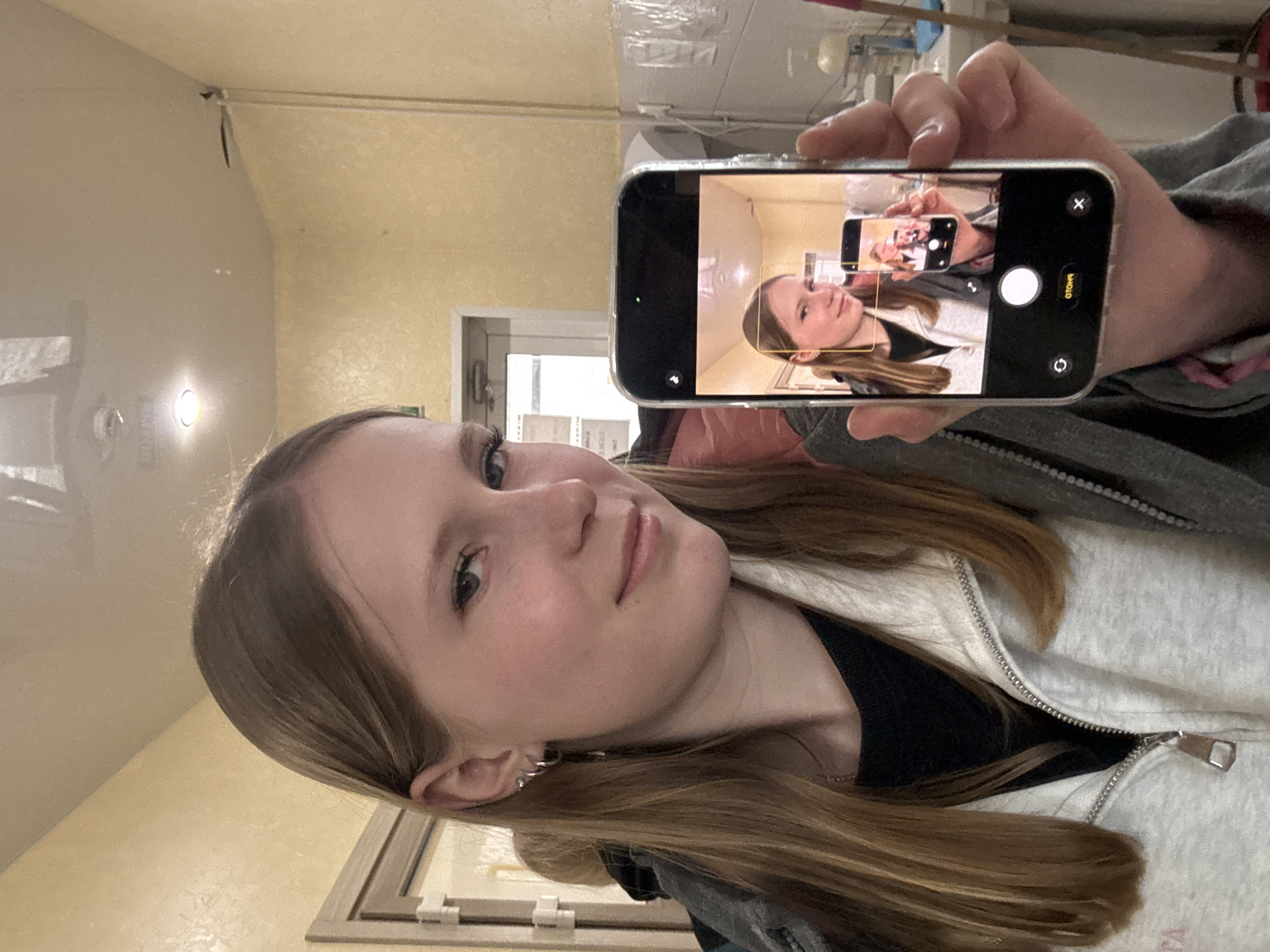 A young woman is taking a selfie while holding a phone that is displaying a selfie of her taking a selfie. She has light brown hair and is wearing a gray zip-up jacket over a black top. The background appears to be an indoor space with yellow walls and a door leading to another room. A light fixture is visible on the wall. The photo does not provide sufficient information to determine the location in Gory, Russia.