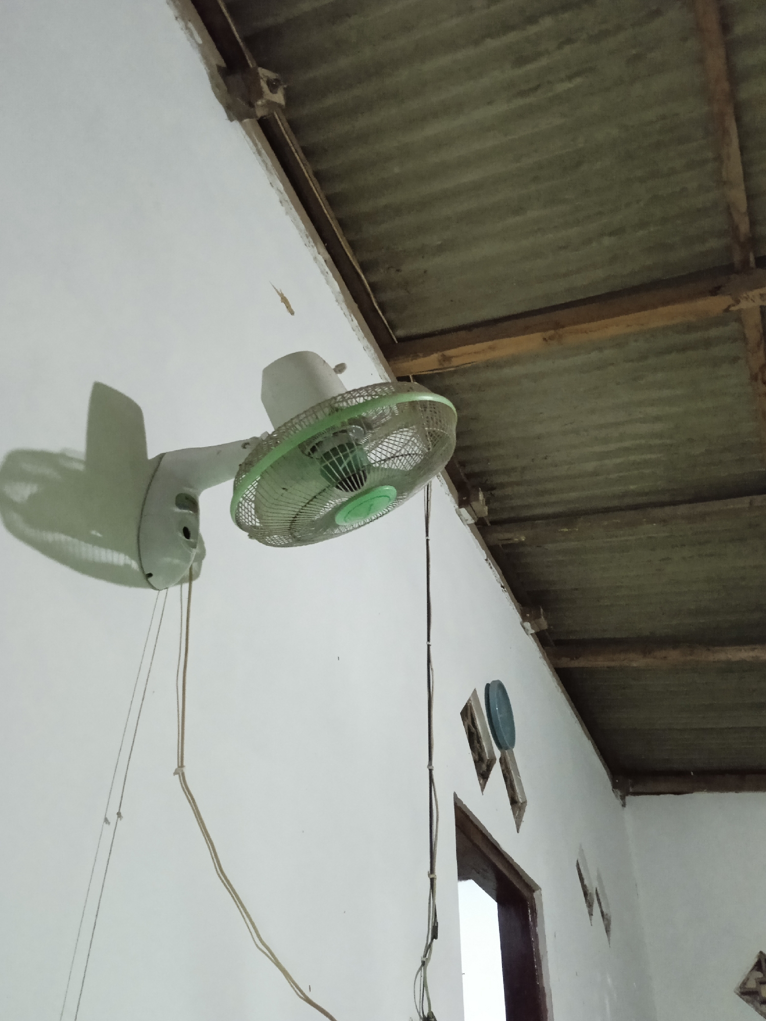 In the city of Kelurahan, Indonesia, a white wall fan is mounted on a white wall. The fan has a green grill and a white body. The fan is angled downwards, and its shadow is cast on the wall. Above the wall, a corrugated metal roof is supported by wooden beams. There are some ventilation openings in the wall near a doorway. A small lizard is visible on the wall. There are also some wires hanging down from the fan and along the wall. The scene appears to be an interior space.