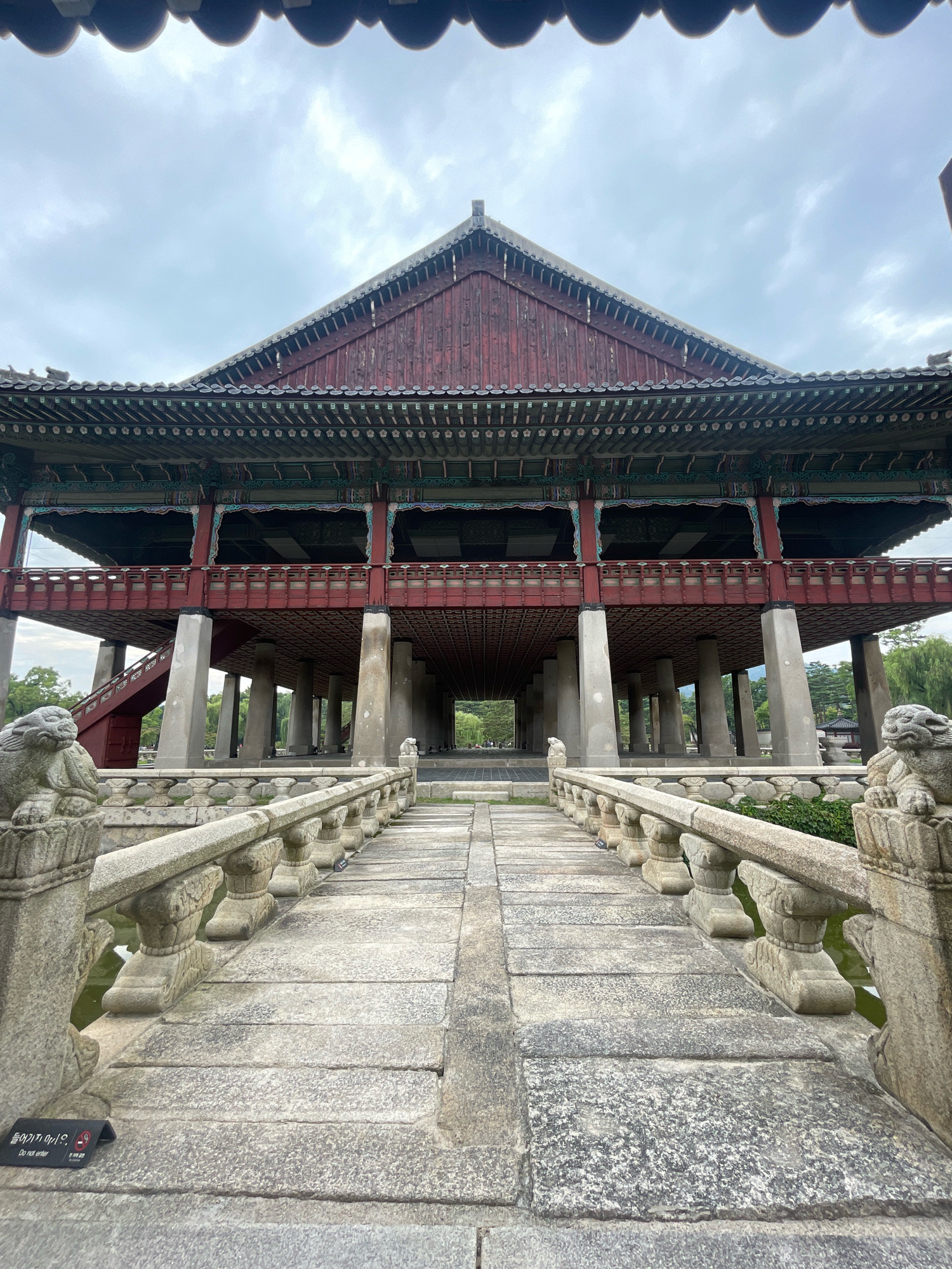 The image shows a historic Korean pavilion, likely in Seoul, South Korea. The pavilion is a multi-story wooden structure with a distinctive tiled roof, painted in deep reds and ornate decorative elements. It is supported by numerous stone pillars, creating an open, airy space underneath. A stone bridge with carved railings and guardian lion statues leads towards the pavilion, spanning a body of water. The bridge and surrounding area are paved with large stone slabs. In the foreground, a small black sign with Korean text and the English words "Do not enter" is visible on the bridge. The sky is overcast with a few faint clouds. The environment appears to be a park or a historical site with trees and greenery visible in the background. There are no people visible in the immediate foreground or on the bridge, though distant figures can be seen walking in the background within the pavilion's grounds.