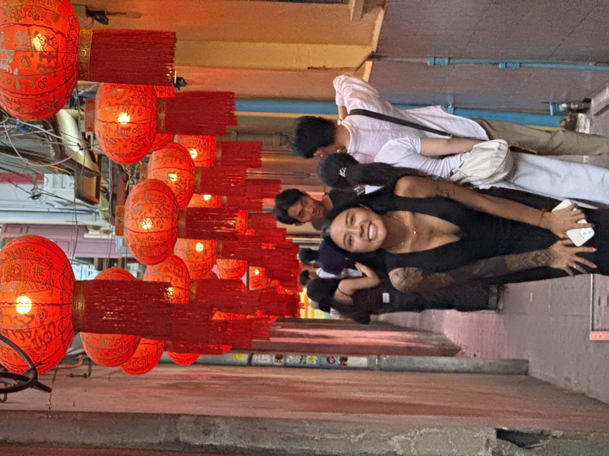 The image depicts a narrow alleyway in Bangkok, Thailand, illuminated by numerous red Chinese lanterns hanging overhead. The lanterns are ornately decorated and emit a warm glow, casting shadows on the surrounding buildings. In the foreground, a young woman with dark hair and tattoos on her arms is smiling broadly at the camera. She is wearing a black dress and holding a smartphone. Behind her, other individuals are walking down the alley, their faces partially obscured. The alley walls are a mix of weathered brick and painted surfaces, with some graffiti visible. The scene conveys a vibrant and lively atmosphere, typical of a bustling street in Bangkok.
