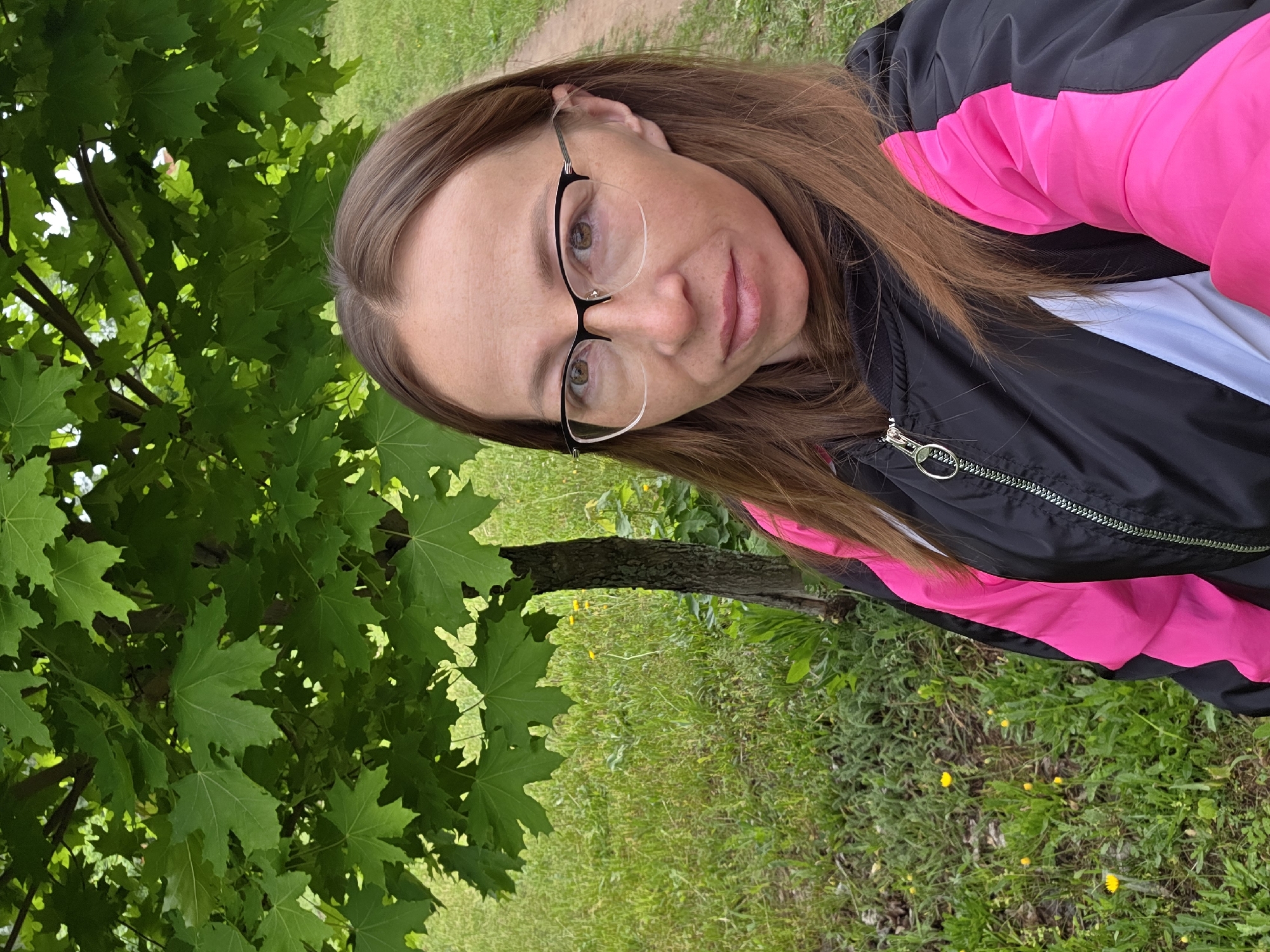 A woman with light brown hair and glasses stands in a grassy, wooded area in the city of Chaikovsky, Russia. She is wearing a black jacket with pink accents over a white shirt. A tree with green leaves is to the left of her face. The ground is covered in grass and some yellow flowers. A dirt path is visible in the background.