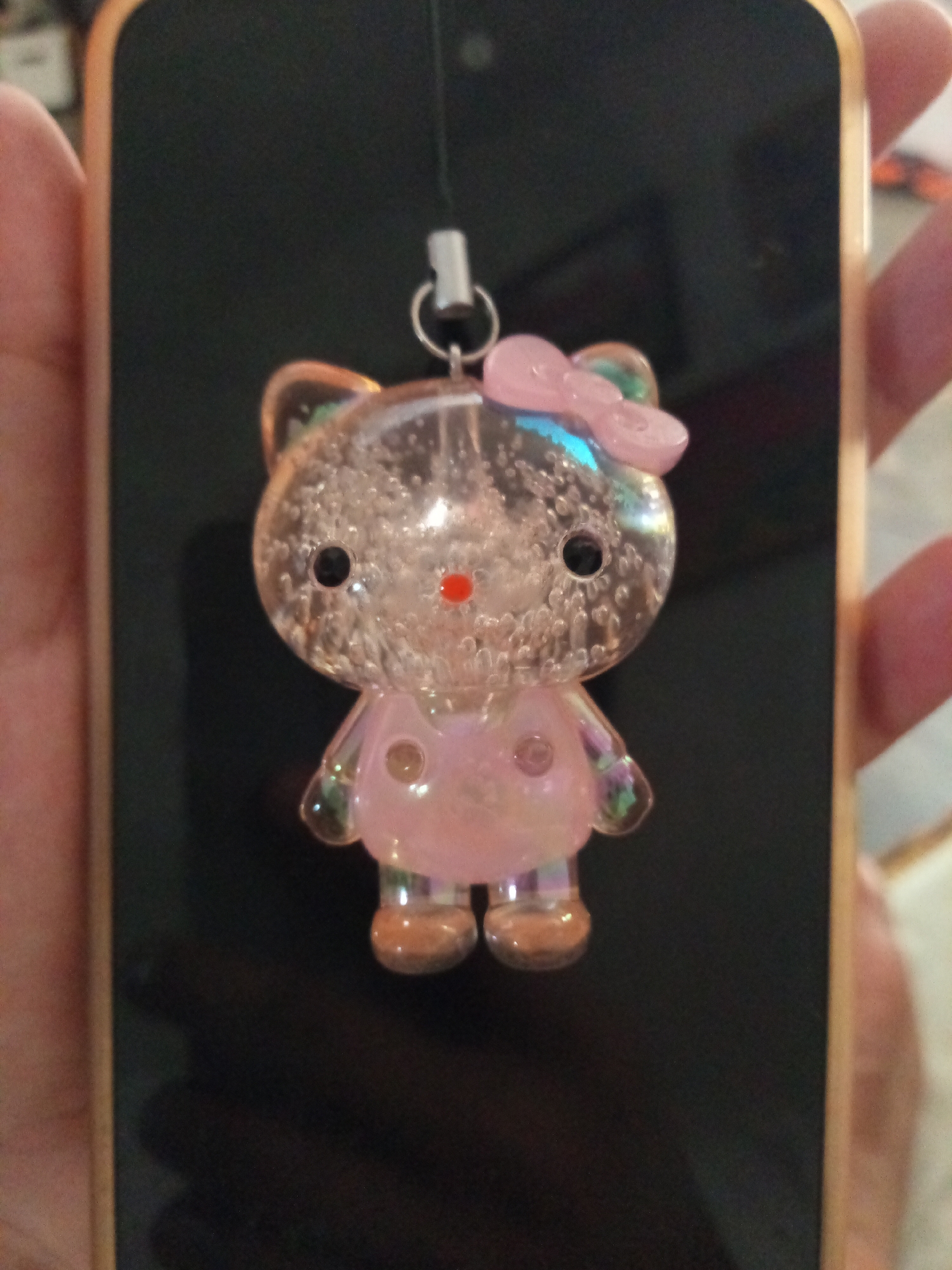 A close-up shot shows a Hello Kitty charm hanging from a phone held in someone's hand. The charm is clear with glitter-like particles inside, featuring a pink bow and a red nose. The phone has a gold-colored case and a black screen. The person's fingers are visible on the side of the phone. The location is unspecified, but the query mentions Davao City, Philippines, which is not discernable from the image itself.