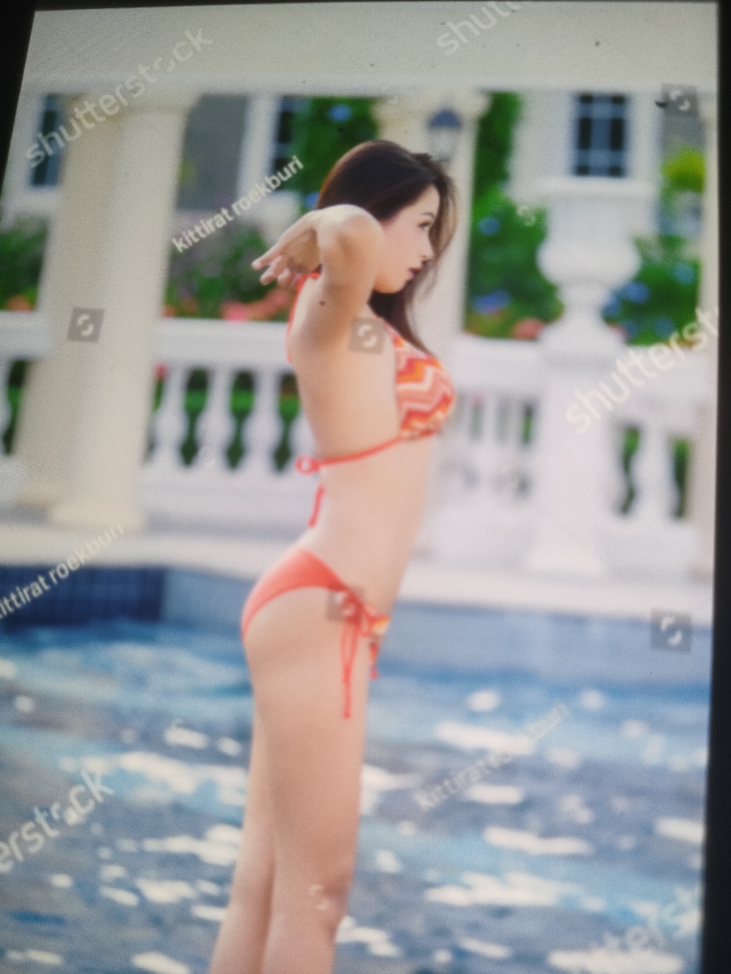 A medium shot captures a woman in a coral bikini standing in a body of water, possibly a pool. She is facing left, with her arms behind her head, stretching her upper body. Her hair is dark and falls down her back. The background includes a white railing, green foliage, and a building with windows. The water has a rippling effect. There are several watermarks present, including "shutterstock" and "kittirat roekburi." The location is not specified as Padang Dalom, Indonesia.
