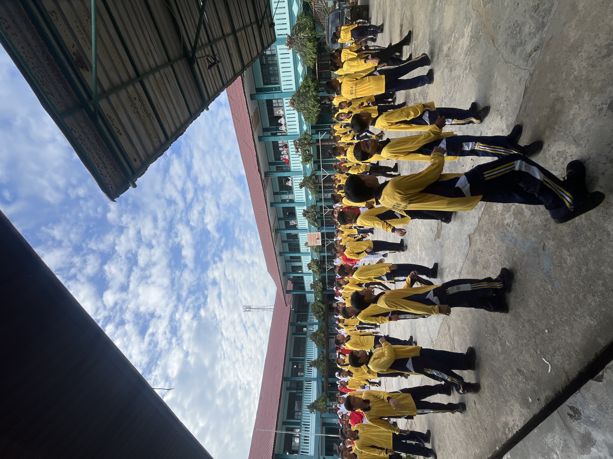 The image shows a group of male students in yellow shirts and dark pants participating in an outdoor activity, likely a school exercise or assembly, in the courtyard of a school in Lhokseumawe, Indonesia. The school building, with its teal and red facade and multiple levels, forms the backdrop. The sky above is partly cloudy with a clear blue expanse. The ground is paved with concrete. The students are arranged in rows, facing the same direction, and appear to be in motion. Some students are more clearly visible than others, with details on their uniforms and facial expressions varying due to their positions. The presence of covered structures, possibly awnings or roofs, frames the top and right side of the image. The overall scene depicts a typical school day activity in an Indonesian educational setting.
