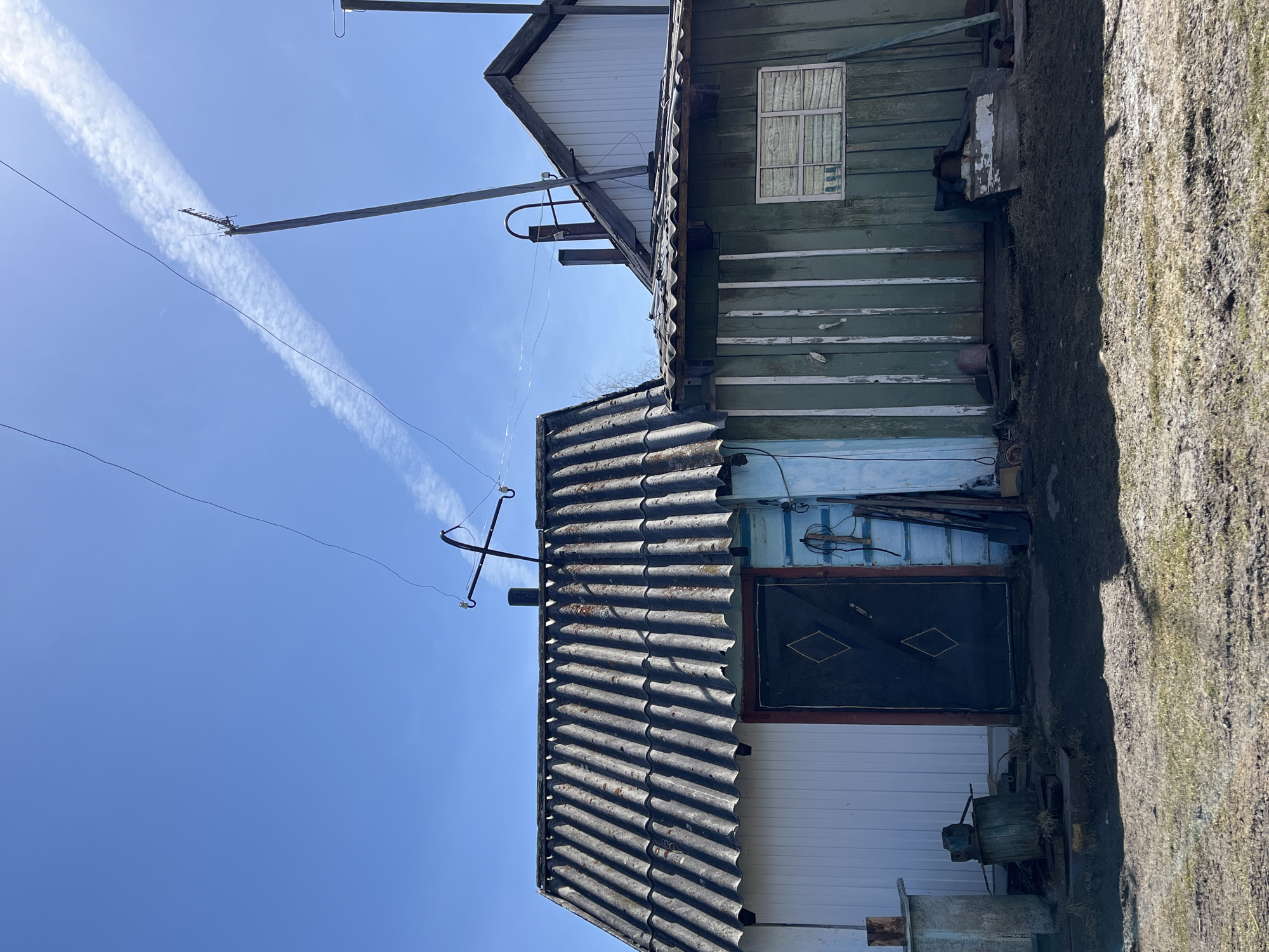 A low-angle, rotated outdoor shot depicts a multi-level building under a bright blue sky marked by a contrail.
The building features white horizontal siding at its base, transitioning to a blue-painted wall section topped by a gray, corrugated roof with an antenna. A dark door with diamond-shaped embellishments sits in the blue wall. Above, the structure shifts to green painted wood, and is topped by another section with white siding. The building is weathered, showing signs of age and disrepair.
Power lines and utility poles are visible against the sky. A patch of weedy ground borders the base of the building.
The location is Верхняя Уря, Russia, as this information was provided to me.