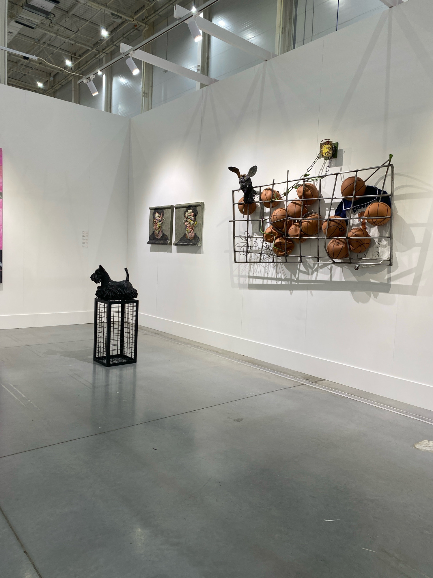 The image shows an art exhibition in a large, well-lit indoor space. The walls are painted white, and the floor is a polished concrete surface. The ceiling features industrial lighting fixtures.
In the foreground, on the left side of the image, is a black sculpture of a Scottie dog sitting on top of a black, grid-like pedestal.
To the right of the dog sculpture, two framed artworks are hung on the wall. They appear to be portraits with somewhat abstract or distorted features.
Further to the right, a large, metal grid structure is mounted on the wall. This structure contains numerous basketballs and a dark blue garment that looks like a basketball jersey. A dark, animal-like sculpture with large ears is attached to the top left of the grid. A chain connects this sculpture to a yellow object mounted on the wall above the grid.
The overall impression is that of a contemporary art exhibition, with a mix of sculpture and wall-mounted art.