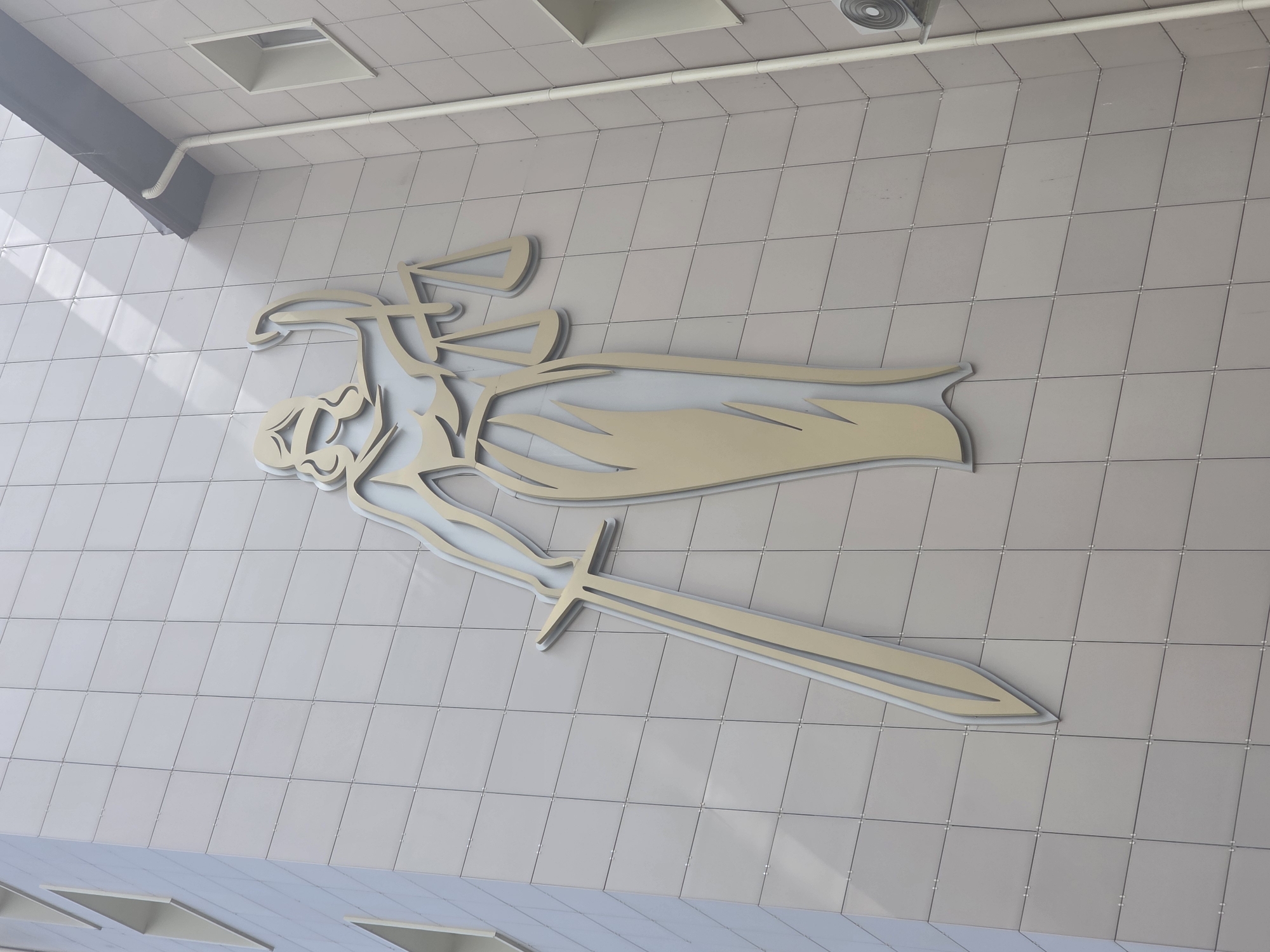 A large, stylized metal sculpture is affixed to the exterior wall of a building. The sculpture depicts a female figure holding a sword and scales of justice, likely representing the legal profession. The sculpture is gold in color with a silver outline. The building has a facade of light gray tiles. The image was captured in Krasnodar, Russia.