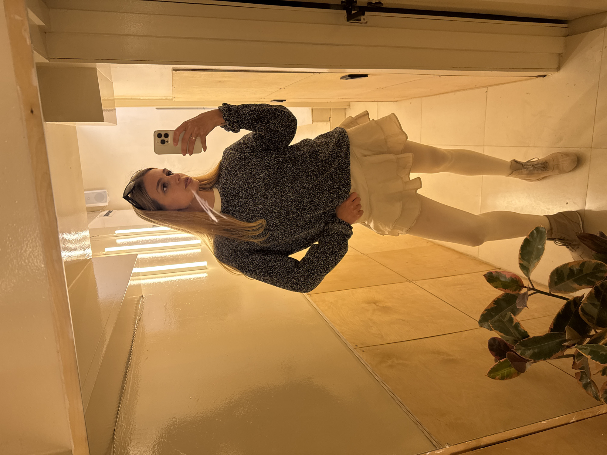 A full-body mirror selfie captures a woman posing in an indoor setting. The image, originally rotated, shows the woman standing upright, holding a light-colored smartphone with a triple camera array in her right hand, pointed towards the mirror. She has long blonde hair and is wearing dark sunglasses pushed up onto her head, with "CATWIE" visible on the frame. Her outfit consists of a dark gray speckled long-sleeved sweater, a light-colored ruffled mini skirt, light-colored tights or leggings, and light-colored lace-up boots.
The background, reflected in the mirror, reveals an interior space with light-colored, possibly glossy, walls and a floor composed of light-toned tiles or wooden panels. Linear light fixtures, likely fluorescent or LED strips, are reflected in the upper left portion of the image. In the lower right corner, a plant with distinctive green and reddish-brown variegated leaves is visible. The lighting is artificial and casts a warm, yellowish hue over the scene. The woman's expression is neutral as she focuses on taking the photograph.
