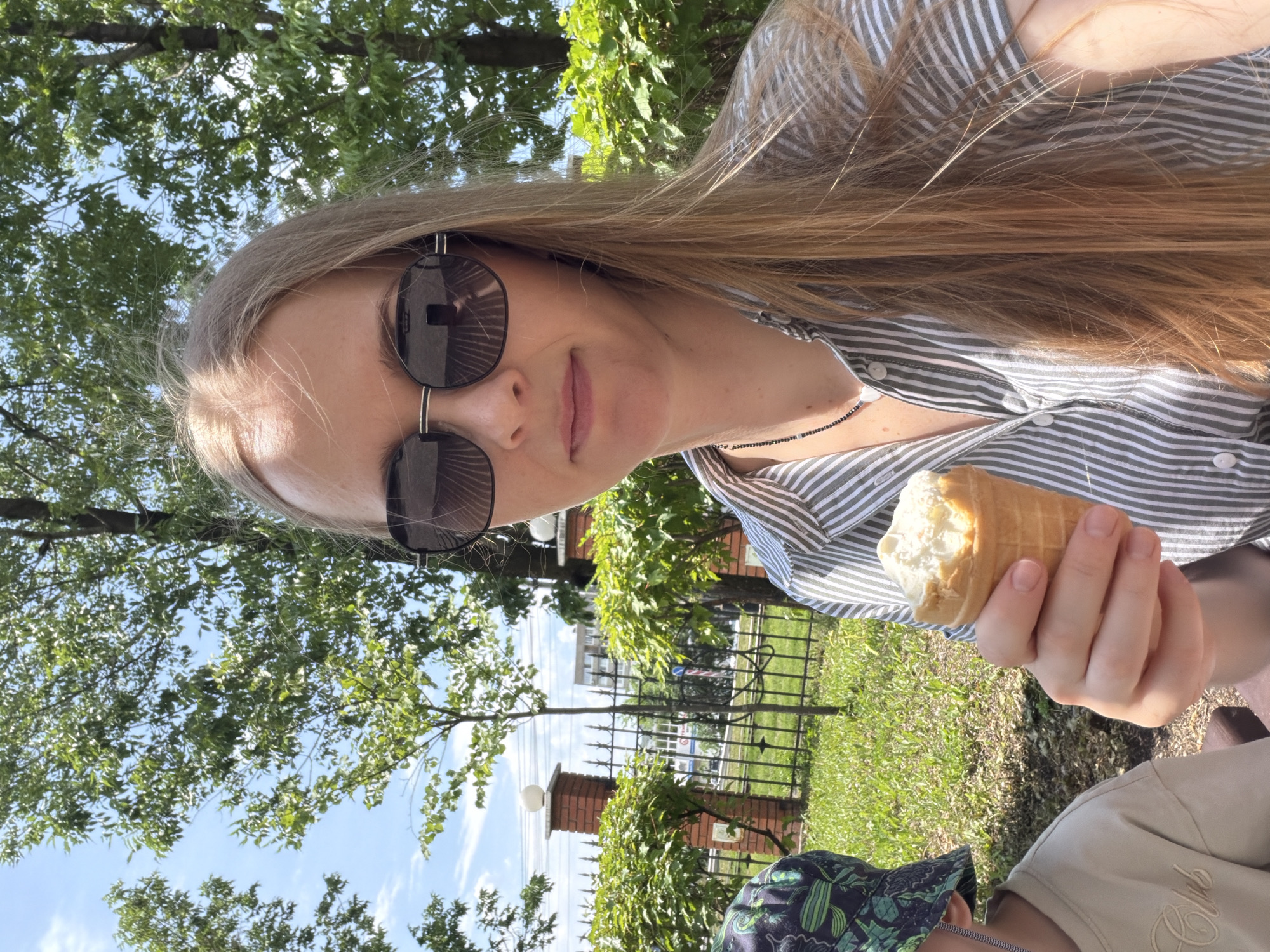A young woman with long blonde hair, wearing large black sunglasses and a striped shirt, holds a small ice cream cone in her right hand. She is taking a selfie outdoors, with a child partially visible on her left. The child wears a green and black patterned hat and a beige t-shirt. They are seated in a park-like setting with green grass, trees, and a black metal fence in the background. Behind the fence, there is a brick pillar with a white ball ornament, and further back, buildings are visible. The sky is blue with scattered clouds. The image appears to have been taken in the city of Khabarovsk, Russia, possibly in a park or public garden.