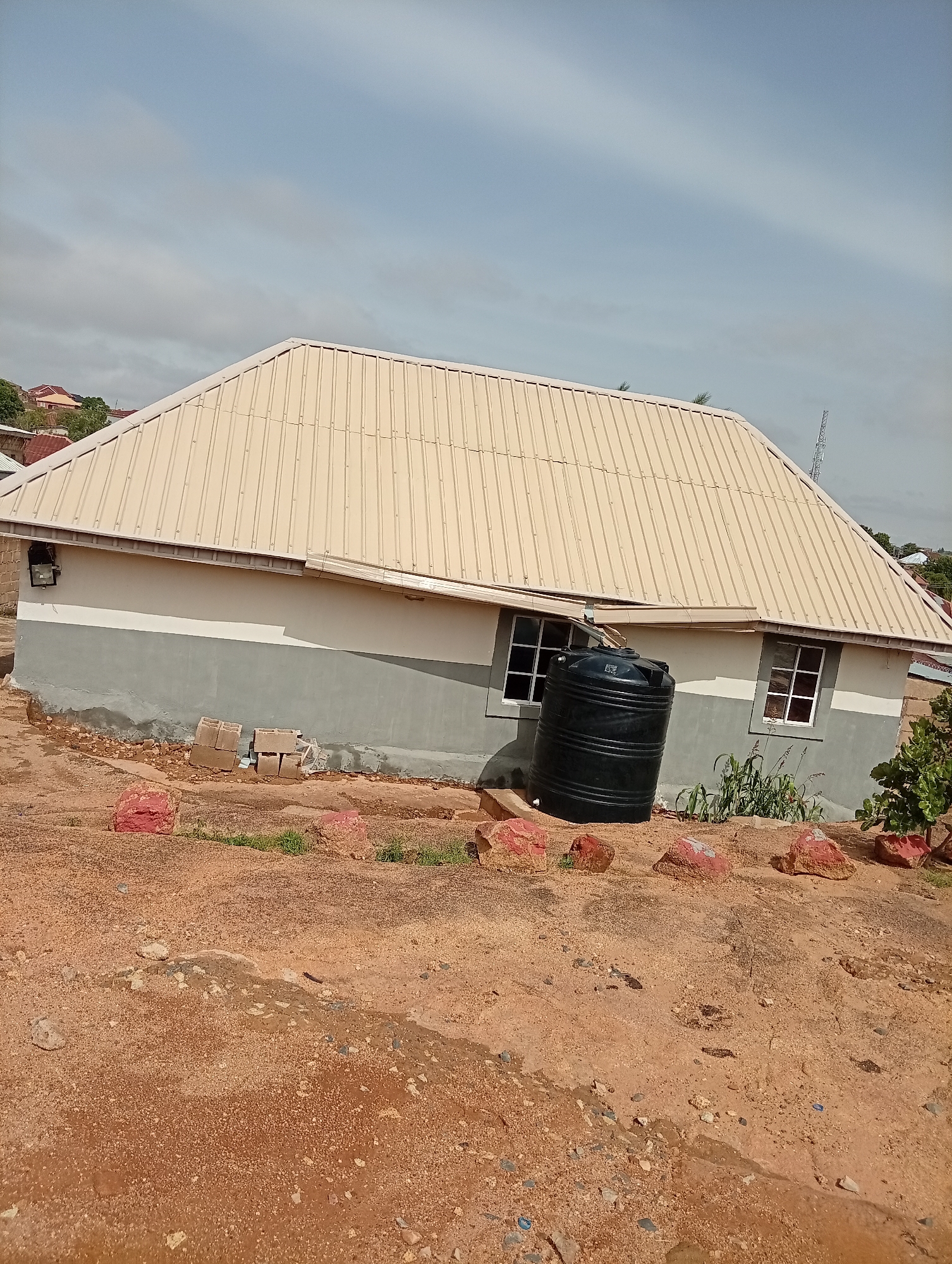 The image shows a single-story building with a light beige metal roof and gray and white walls, located in Jos, Nigeria. The house has at least two visible windows with white frames. A large black water tank stands outside one of the windows. The ground is reddish-brown soil with several red rocks scattered around the base of the house. In the background, other buildings and a communication tower are visible. The sky is light blue with scattered clouds. There are some plants and small bushes near the house. Some construction materials, such as concrete blocks, are visible near one side of the house. The overall impression is that the house is new or recently constructed.
