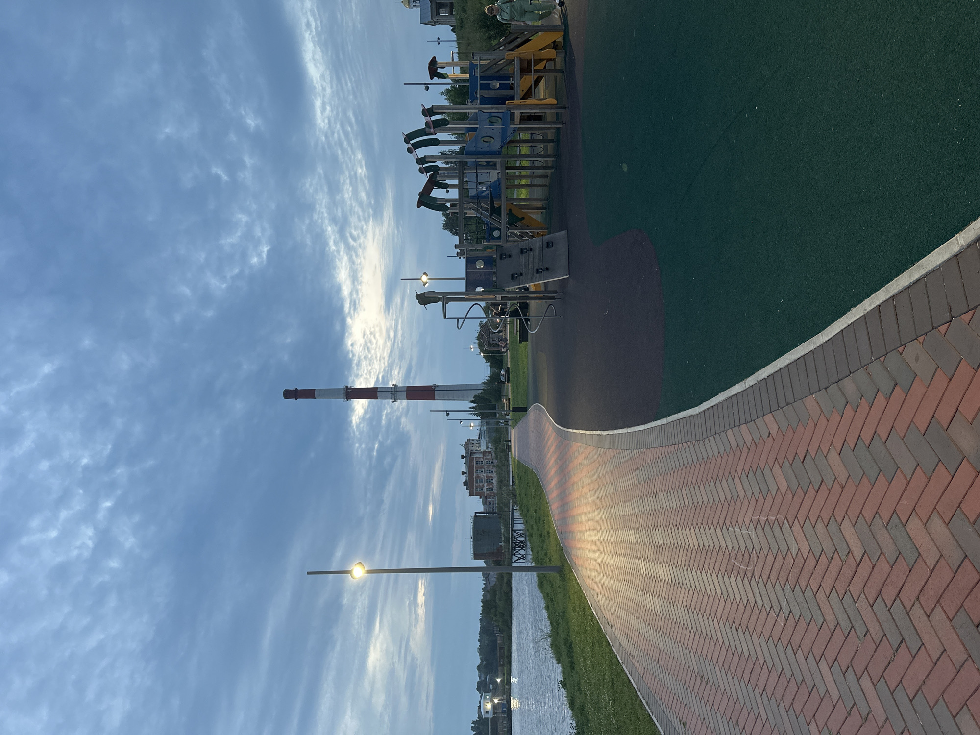 The image depicts a waterfront park area in Lysva, Russia, under an overcast sky. A red and white striped smokestack is visible in the background, along with several industrial and residential buildings. A paved walkway, laid with red and grey bricks in a herringbone pattern, curves along the waterfront. The walkway is illuminated by a lamppost. Adjacent to the walkway is a children's playground with slides, climbing structures, and swings, set on a dark green, shock-absorbent surface. A woman in a light green tracksuit is walking away from the playground on the right side of the image. The water body to the left appears calm, reflecting the sky and some distant lights. The overall scene suggests a public recreational space in an industrial town, likely captured during twilight or early evening.
