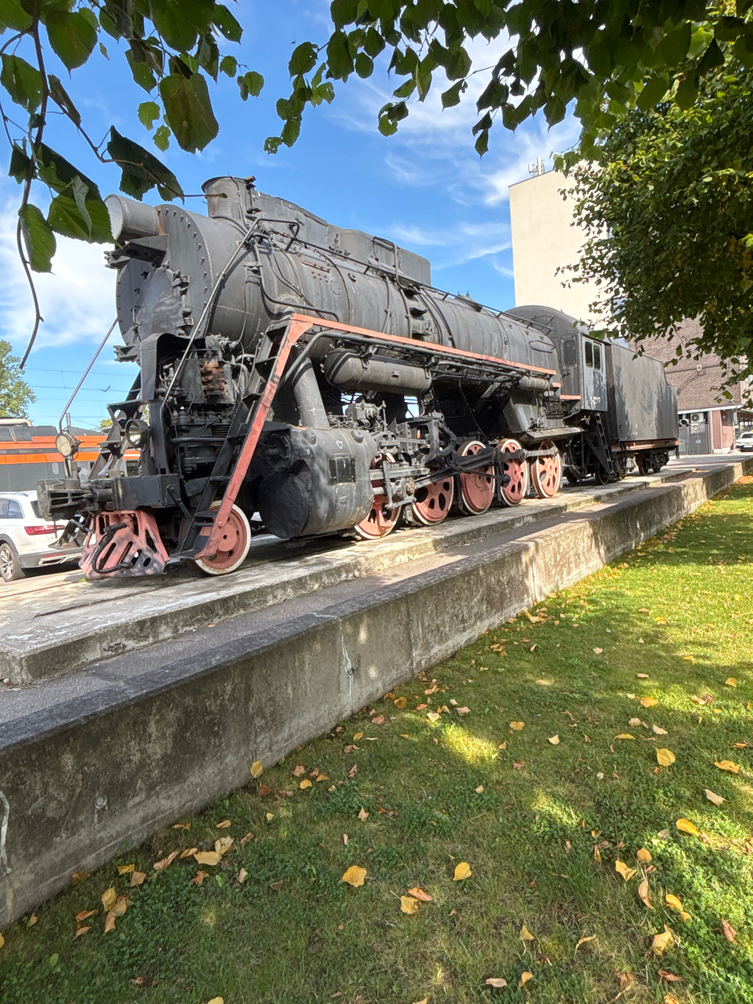 An old, black steam locomotive is displayed on a concrete platform outdoors. The locomotive is viewed from a low angle, with its front facing left. Red accents are visible on the locomotive's wheels, front buffer, and some structural components. Fallen yellow leaves are scattered across the green grass surrounding the platform. In the background, a modern white building is visible, along with trees and a clear blue sky with scattered clouds. A white car is parked to the left of the locomotive. The scene appears to be in a public park or museum. This location is likely the Estonian Railway Museum in Tallinn, Estonia, given its outdoor display of historical locomotives.