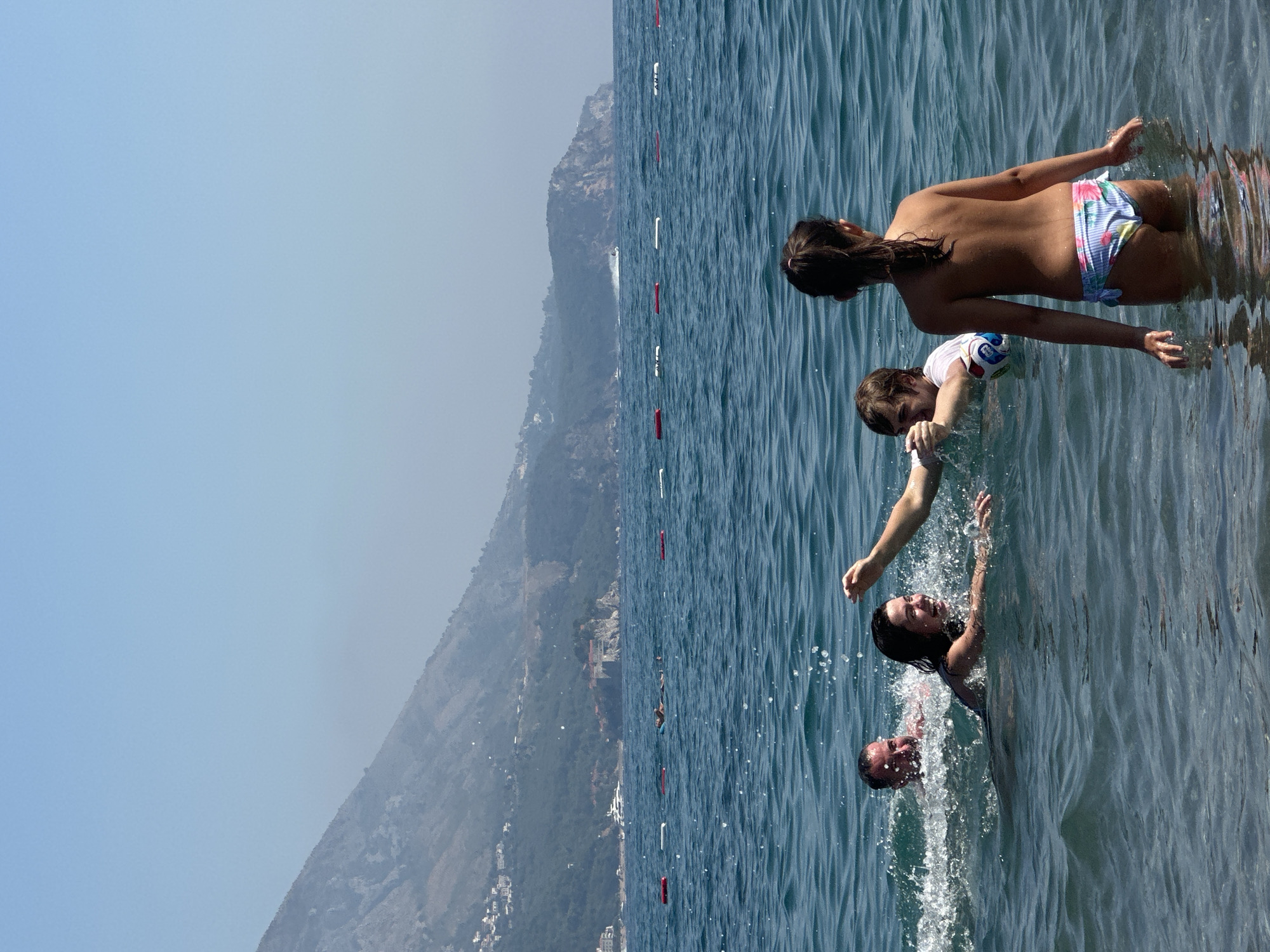 This media file shows a group of people swimming in the ocean. The scene is set against a backdrop of a hazy, mountainous coastline with scattered buildings. The water is a vibrant blue, with gentle waves and sunlight reflecting off the surface. In the foreground, four individuals are visible. One person, wearing a floral bikini, is partially submerged in the water, facing away from the camera. Another person, in a white shirt, is swimming towards the camera, holding a colorful ball. Two more people are in the water, one with their head and shoulders visible, and another submerged. The overall impression is one of a fun and active day at the beach. The time of day appears to be daytime, with clear skies and bright sunlight. The location cues, such as the mountainous coastline and the clear blue water, suggest a Mediterranean or similar coastal environment. The expressions on the faces of the people visible suggest enjoyment and exertion. There is no visible text in the image.
