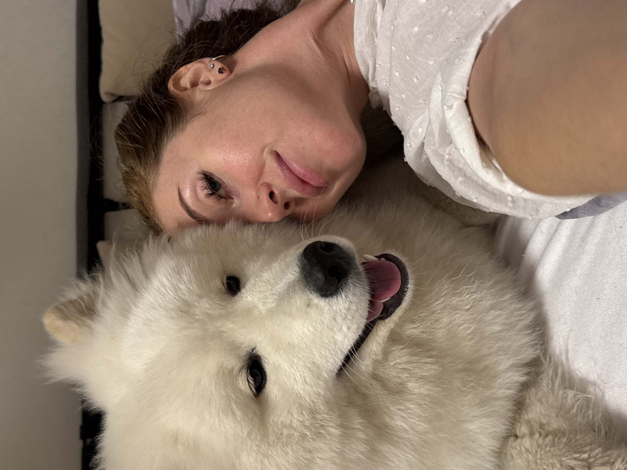 A woman and a large, white Samoyed dog are lying down together. The woman is on the left side of the image, her head resting on the dog's fur. She has light skin, brown hair, and is wearing a white top with small patterns. The dog is positioned below her, with its head close to the camera. The dog's mouth is open, its tongue visible, and its eyes are alert. The background is blurred, suggesting an indoor setting, possibly a bed or a couch. The location is unspecified.