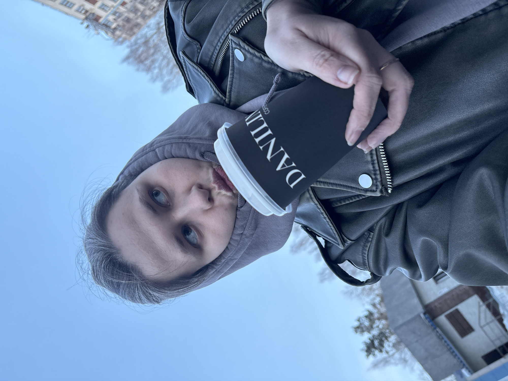 A person, likely female, with light greyish-blonde hair, is depicted in an outdoor, upward-angled selfie-style shot in Chaikovsky, Russia. The individual wears a grey hooded sweatshirt underneath a dark grey or olive-green leather jacket featuring silver zippers and snap buttons. Their face is angled to the right, with eyes looking upwards and to the right, and lips pursed into a pouting expression. The right hand holds a black disposable coffee cup with a white lid, bearing the text "DANILIN COFFEE" in white capital letters. A simple gold-colored ring is visible on the left ring finger.
The background consists of a clear, light blue sky. To the right, a multi-story, light-colored apartment building with visible windows is present, out of focus. To the left, a smaller, light-colored building with a dark-trimmed roof and some visible steps is partially obscured. Bare deciduous trees and some evergreen trees are scattered in the background. Patches of snow are visible on the ground, particularly near the smaller building, indicating cold weather conditions. The scene is consistent with an urban setting, specifically near a "DANILIN COFFEE" establishment, which is located at Lenin Street, 52, Chaikovsky, Perm Krai, Russia.