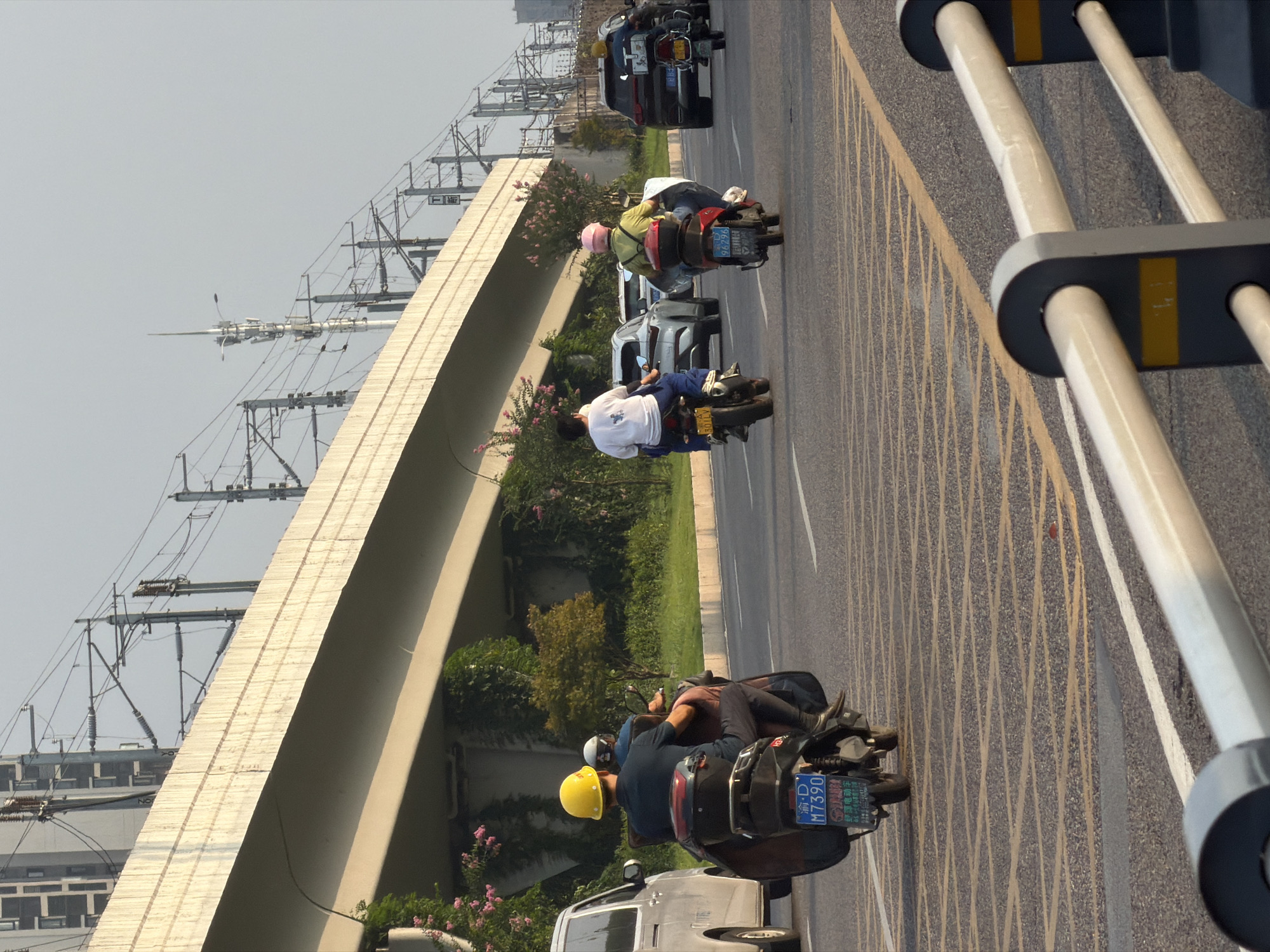 In the city of Jiangjin, China, a road scene unfolds under an overcast sky. A person wearing a yellow hard hat rides a scooter, facing away from the camera and towards the road ahead. Behind them, another person sits on the same scooter. To their left, a white van is parked. In the background, a concrete overpass supports an intricate network of electrical wires and poles, suggesting proximity to a transportation hub. The road is marked with yellow and white lines, indicating multiple lanes of traffic. Further down the road, several other vehicles, including a black SUV and two more scooters, are visible. The general environment appears to be urban, with some greenery and trees lining the sides of the road.