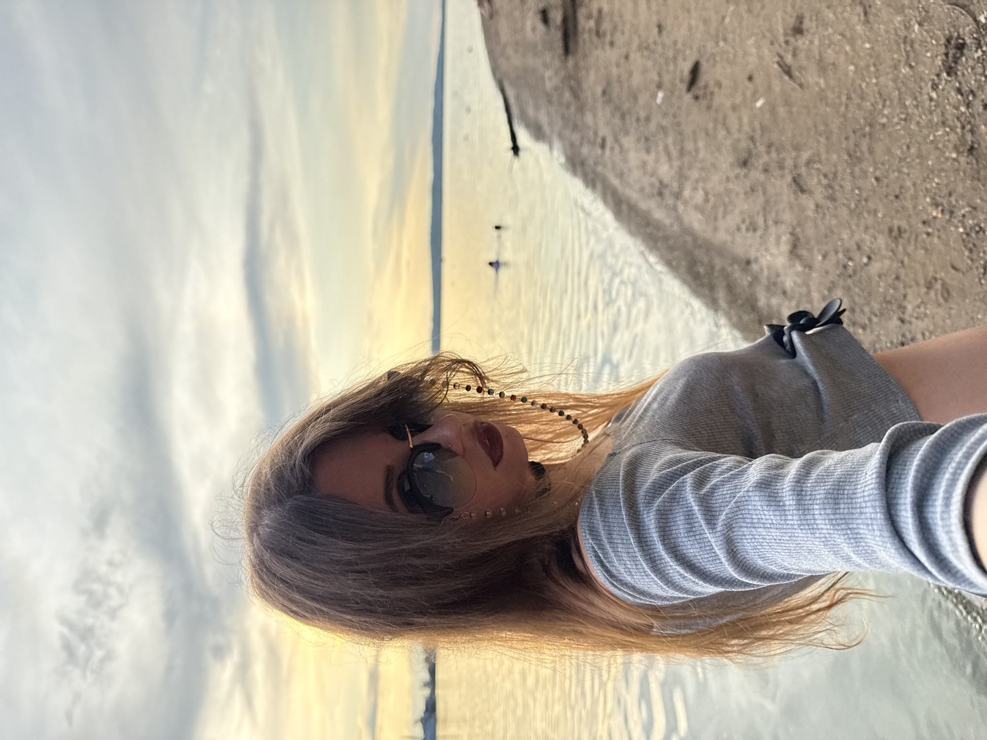 A young woman with long brown hair, wearing oversized sunglasses and a grey off-the-shoulder crop top with long sleeves, stands on a sandy beach. Her hair is styled in loose waves, and she is looking over her right shoulder towards the camera. A beaded chain is attached to her sunglasses. The setting is a beach with calm water stretching out to the horizon under a cloudy sky with hints of yellow from what appears to be a sunset. In the distance, there are a few people in the water and some industrial structures on the far left. The sand on the beach is light brown and appears somewhat disturbed.