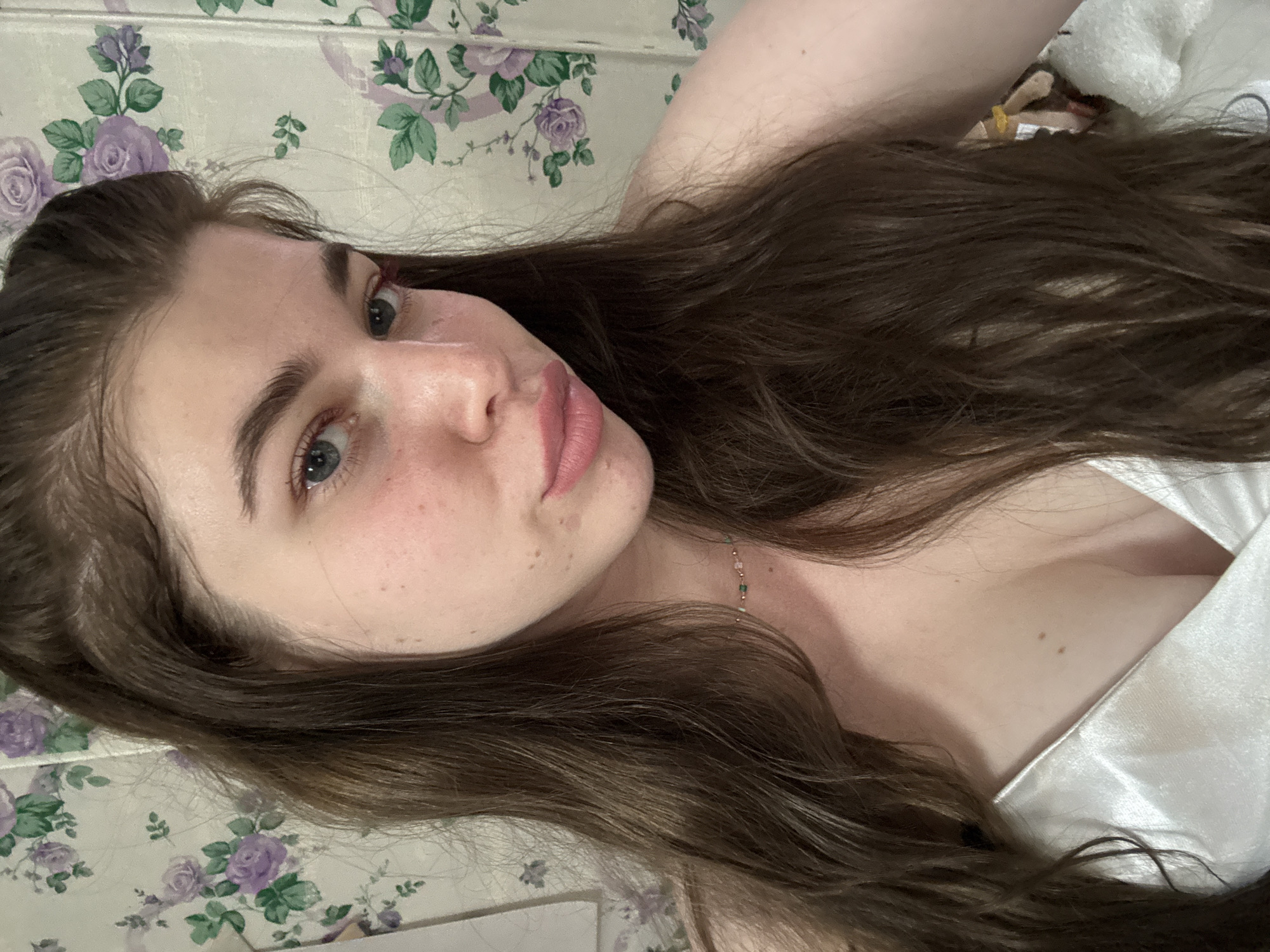 A young woman is lying down, facing the camera. She has long, brown hair, fair skin with freckles, and light-colored eyes. She is wearing a light-colored top. The background is a floral patterned surface. The location is not discernible from the image.