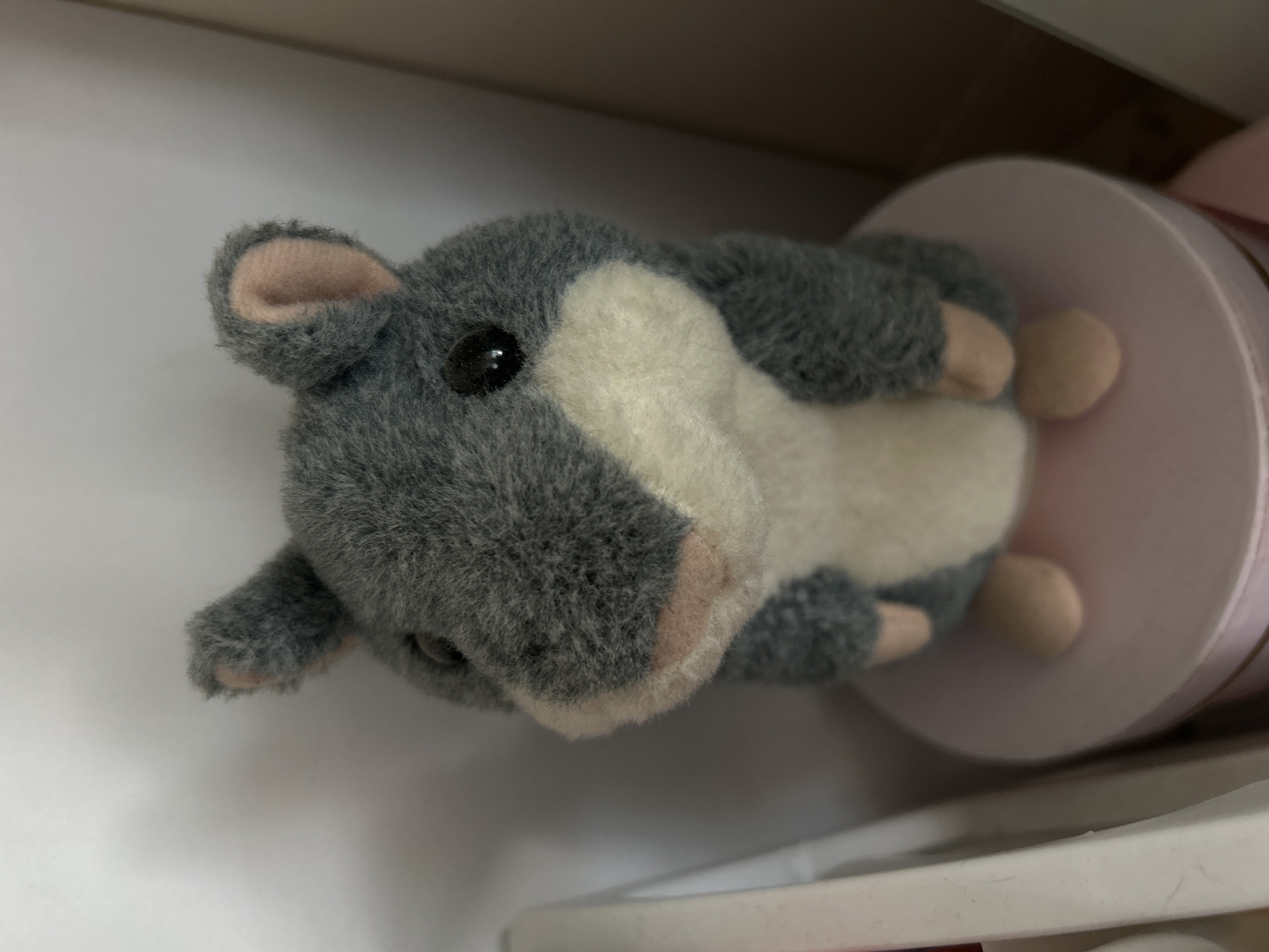 A plush toy resembling a gray and white hamster or guinea pig is captured in a close-up, horizontal shot. The toy features soft, fluffy gray fur covering its back, head, and limbs, complemented by a patch of creamy white fur on its belly and face. Its small, rounded ears have pale pink interiors, and a single glossy black eye is visible on its left side. The snout area is white with a light pink nose.
The stuffed animal is positioned on its back, with its head facing the left of the frame. Its small, beige-colored paws are visible, resting against a light pink cylindrical object located on the right side of the image. The background is indistinct, showing blurred white and light gray surfaces, suggesting an indoor setting such as a shelf or a display. No activity is depicted, as the toy is stationary. There is no visible text in the image.