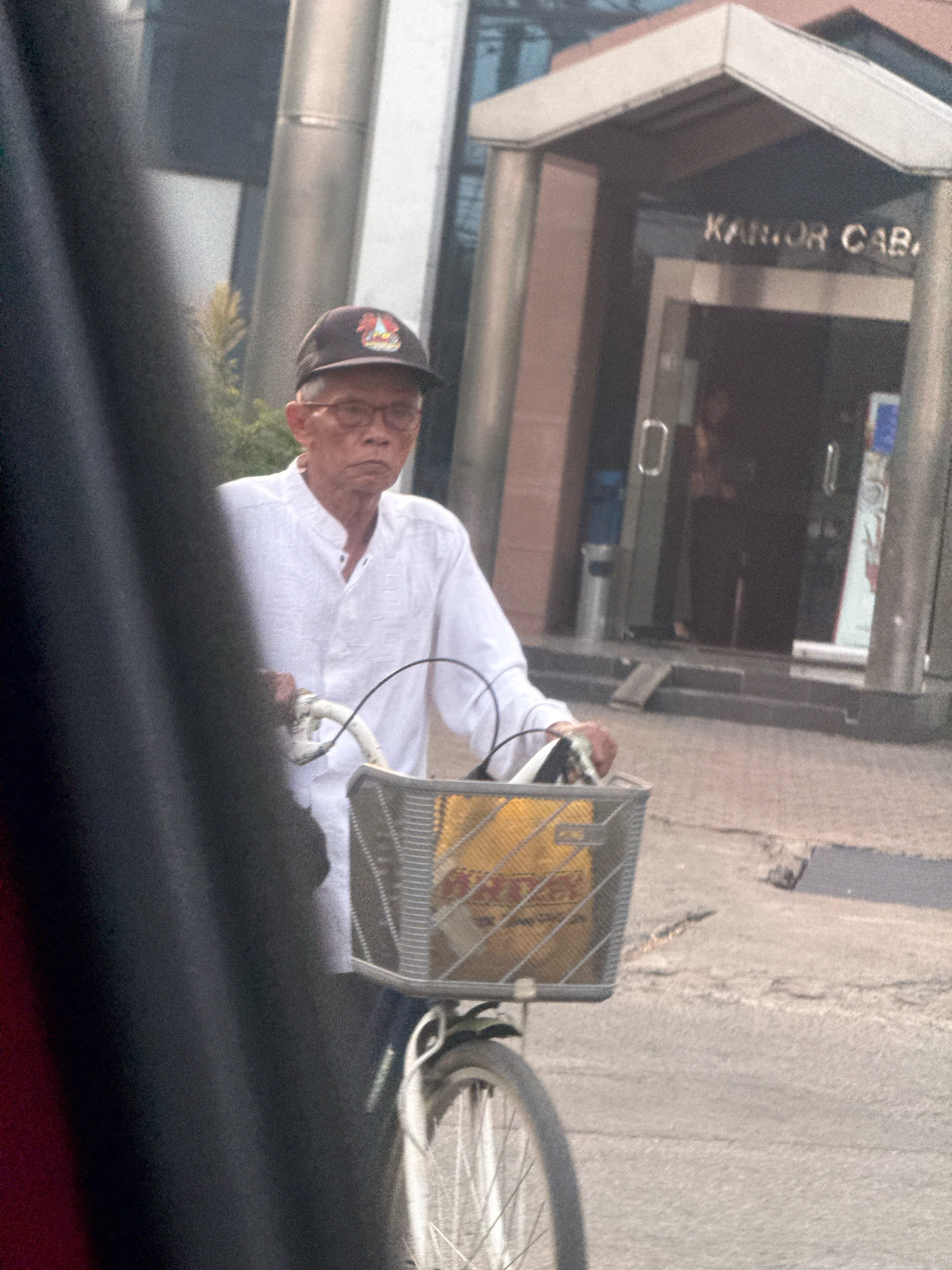 An elderly Asian man wearing glasses and a white, collared, long-sleeved shirt, and a baseball cap is riding a bicycle with a basket in front. The basket contains a yellow object with red lettering. The man is looking straight ahead with a neutral expression. The background shows a building with a sign that reads "KANTOR CAB" in large letters, likely an office building. The scene appears to be on a street in Surabaya, Indonesia, with a blurry dark object on the left side of the frame possibly being a vehicle.