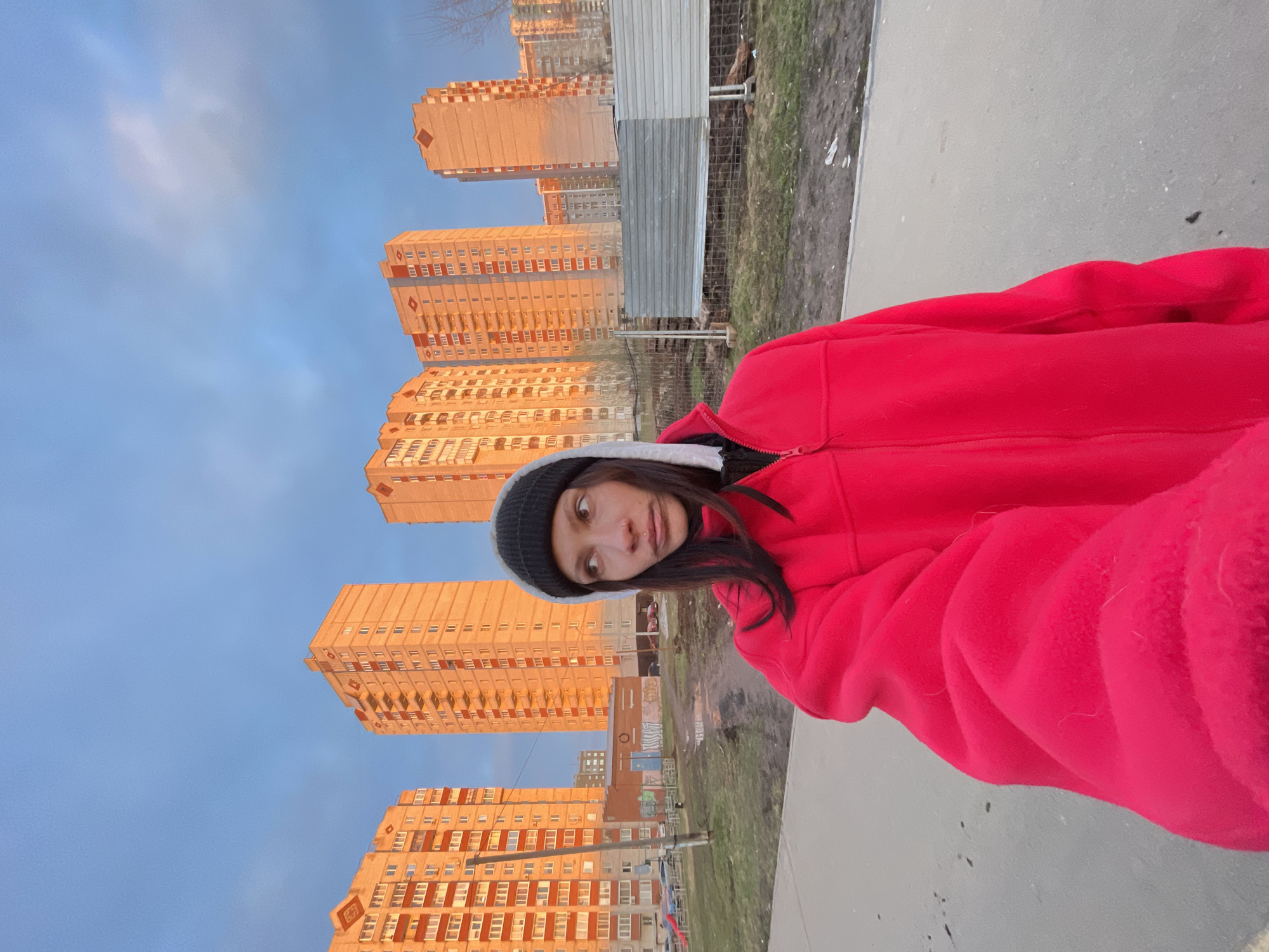 This vertical outdoor portrait captures a young woman in an urban setting, identified as Izhevsk, Russia. The woman, with dark hair and a nose piercing, is positioned on the right side of the frame, looking towards the left with a slightly curious expression. She is wearing a bright red, puffy jacket and a dark beanie hat.
Behind her, dominating the left side of the composition, are several tall, multi-story residential buildings. These buildings, characteristic of post-Soviet architecture, are bathed in warm, golden light, indicating either sunrise or sunset (golden hour). The sky above is a clear, light blue.
In the mid-ground, separating the woman from the buildings, is an area of rough ground featuring patches of dry grass and dirt. A temporary barrier, constructed from corrugated metal sheets and chain-link fencing, runs across this area. The woman appears to be standing on a paved pathway or sidewalk that occupies the foreground.
The image conveys a casual, everyday scene, capturing a moment during a clear, possibly cool, day in an Izhevsk neighborhood. No discernible text is visible in the image.
