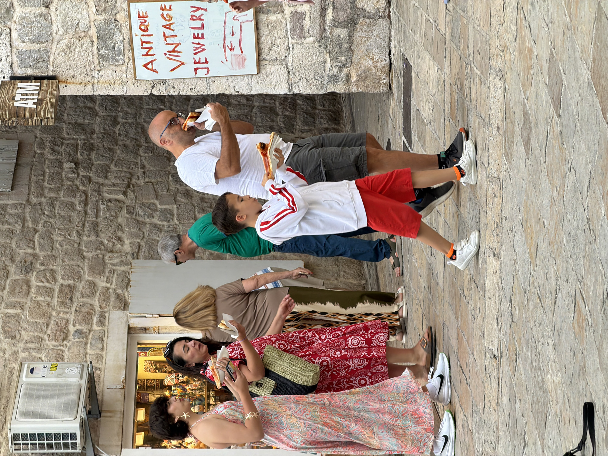 A group of people is walking on a cobblestone street in Budva, Montenegro, near an ATM and a shop selling antique and vintage jewelry. A young boy in a white jacket with red stripes and red shorts is walking towards the camera, holding a slice of pizza. A man in a white t-shirt and gray shorts is walking behind him, also eating pizza. Two women are walking in the opposite direction, one in a pink and white patterned dress and the other in a red dress. A woman with blonde hair wearing a brown shirt and patterned pants is walking past them. The buildings are made of stone, and an air conditioning unit is mounted on the wall of one of them.