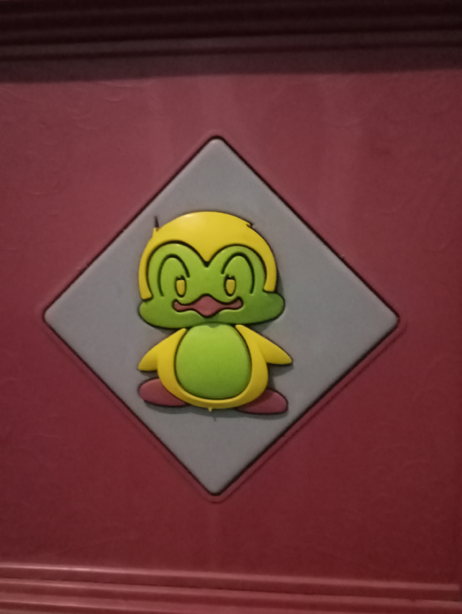 The image shows a close-up of a cartoon character sticker on a red, textured surface. The character is primarily green and yellow, with large eyes and a wide mouth. It appears to be a stylized depiction of a bird or a frog. The sticker is affixed to a diamond-shaped gray background, which is itself set against the red surface. The red surface has horizontal ridges at the bottom, suggesting it might be part of a piece of furniture or a decorative panel. The lighting is somewhat dim, casting soft shadows. There are no people or other identifiable objects in the frame. The location is stated to be Kecamatan Haurgeulis, Indonesia, but there are no visual cues in the image to confirm or deny this.