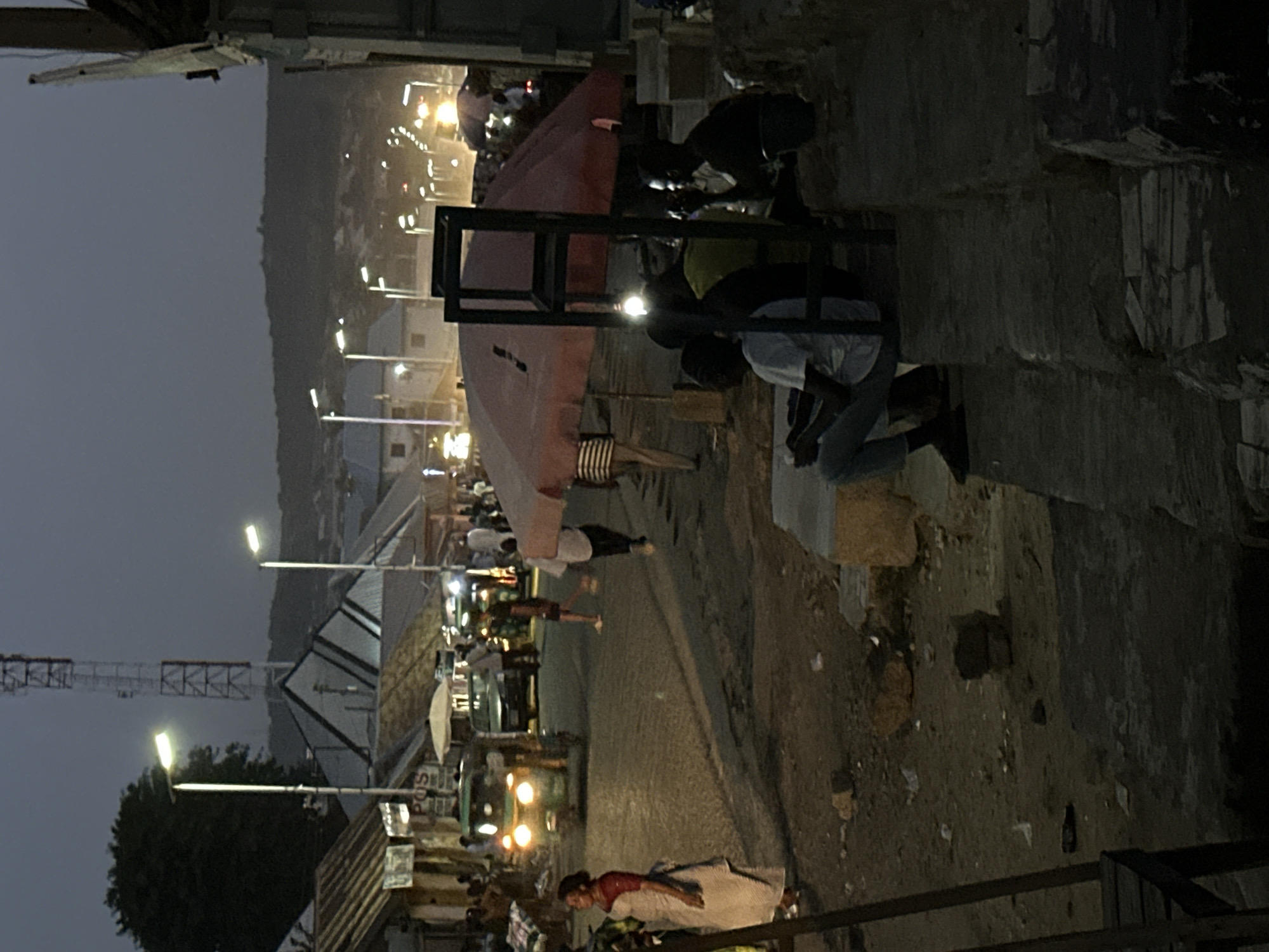 This image depicts a street scene in Jos, Nigeria, during what appears to be twilight or early evening. Streetlights illuminate the road, which is lined with buildings and populated with people. Individuals can be seen walking, some standing near stalls under a red awning. Some appear to be praying. Vehicles, including what appear to be automobiles and auto rickshaws, are visible on the road. A communications tower is visible in the background. The overall environment appears somewhat urban and potentially busy with activity.