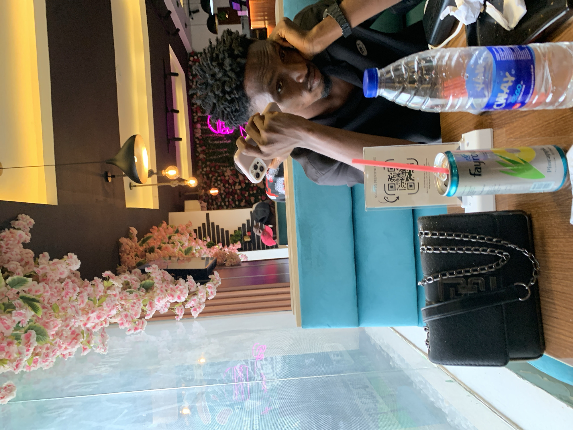 The scene is an indoor shot of a person sitting at a table in what appears to be a restaurant or café.
The person is holding a smartphone, possibly taking a photo or using it. On the table, there is a can of pineapple drink with a straw, a water bottle, a menu, and a small black purse with a silver chain.
The environment is decorated with flowers. There is striped wallpaper and other decorative elements in the background. The restaurant appears to be located in Adankolo, Nigeria.