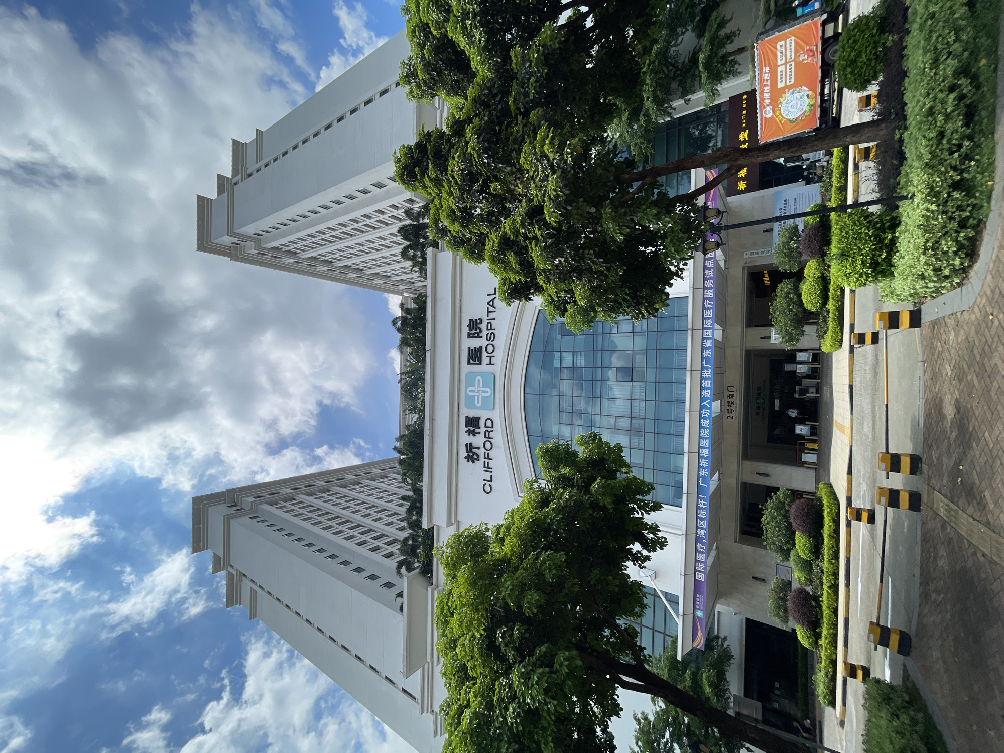 A low-angle, wide shot captures the imposing facade of Clifford Hospital under a partly cloudy sky in Panyu District, China. The building features two towering, white, modern skyscrapers symmetrically flanking a central, glass-fronted entrance. Lush green trees and manicured shrubbery are strategically placed in front of the building. The hospital's name, "CLIFFORD HOSPITAL," is clearly visible in English above the entrance, accompanied by Chinese characters and a blue cross symbol. A vibrant banner, displaying Chinese text and graphics, is draped across the upper part of the entrance area. Yellow and black striped barriers are positioned on the road leading to the hospital. A truck with an orange advertisement is parked to the right of the entrance. The overall scene conveys a sense of modern healthcare infrastructure and a bright, daytime setting.