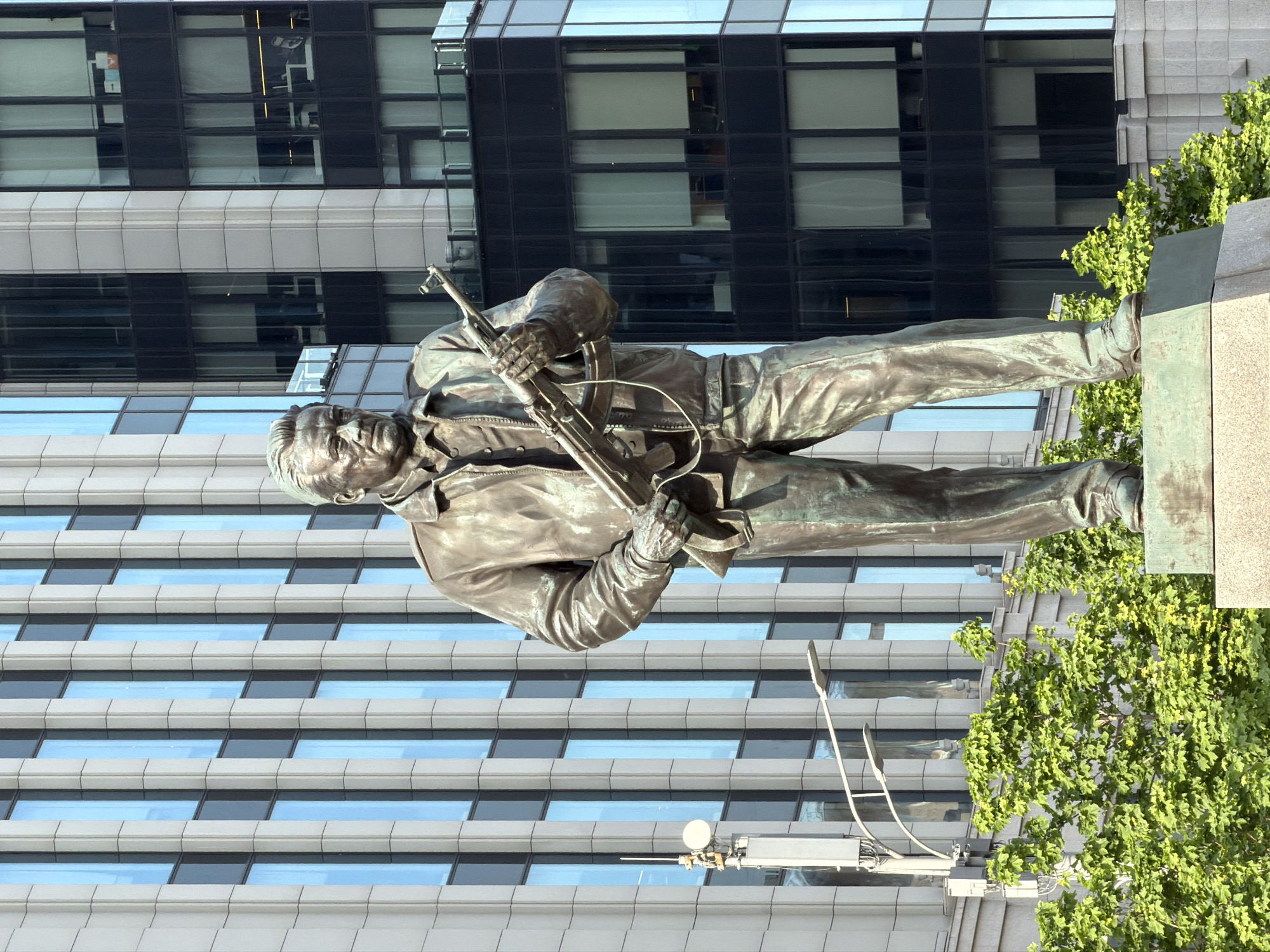 The image shows a statue in front of a building in Moscow, Russia. The statue is of a man holding a rifle, likely a monument to a military figure. The surrounding environment includes greenery and a modern building facade. A street light is also visible.