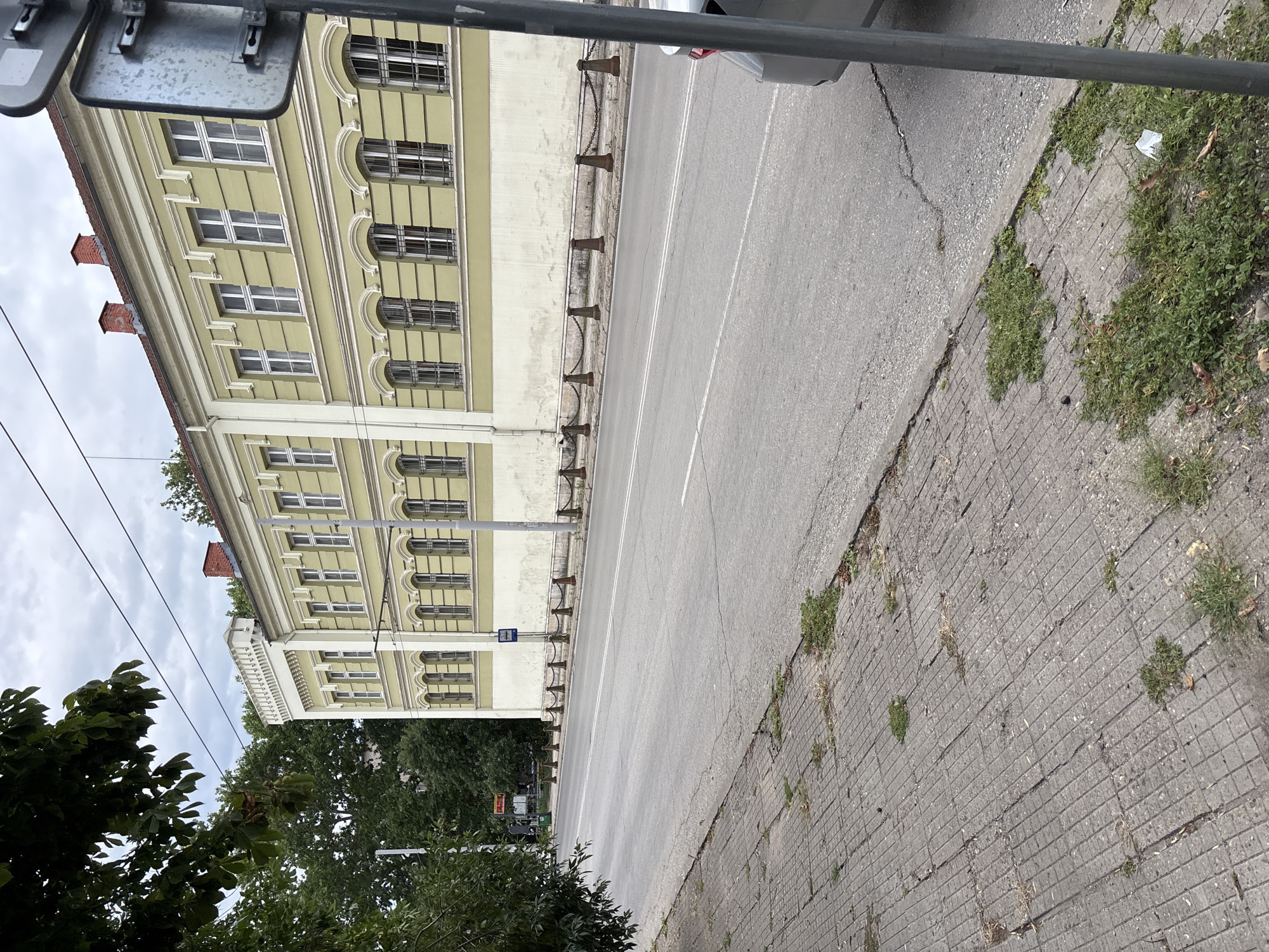 The image shows a street scene in Pleven, Bulgaria, with a two-story building on the right and a sidewalk on the left. The building, painted in a light yellow, features multiple windows with arched tops on the lower floor and rectangular windows on the upper floor. A red-tiled roof with chimneys is visible at the top. A chain fence with bollards separates the sidewalk from the street. The sidewalk is paved with square concrete tiles and has patches of grass growing through the cracks. The street is asphalted with double white lines marking the lanes. A grey car is partially visible on the right side of the road, and a street sign on a metal pole stands near the sidewalk. Trees with green foliage are on the left side of the image, partially obscuring the view. The sky is overcast with grey clouds.