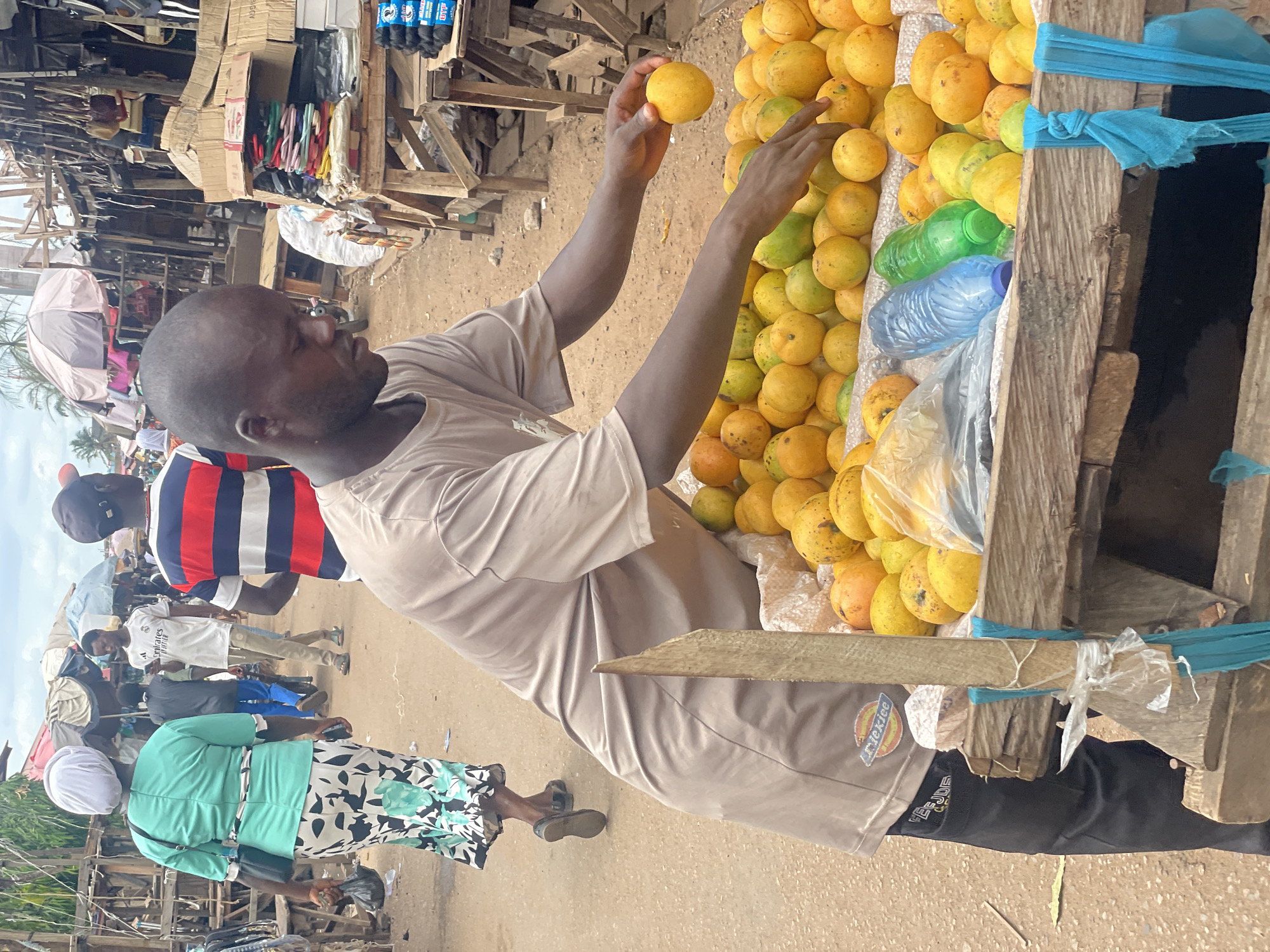 A man stands at a wooden stall piled high with yellow mangoes in Lokoja, Nigeria. He wears a light brown t-shirt and dark pants, his arms raised to select a mango. To the left, people pass by on a dirt road, including a woman in a green top and floral skirt. In the background, other stalls are visible, creating a bustling marketplace environment. Bottles of water sit amongst the fruit, and blue fabric adorns the stall.