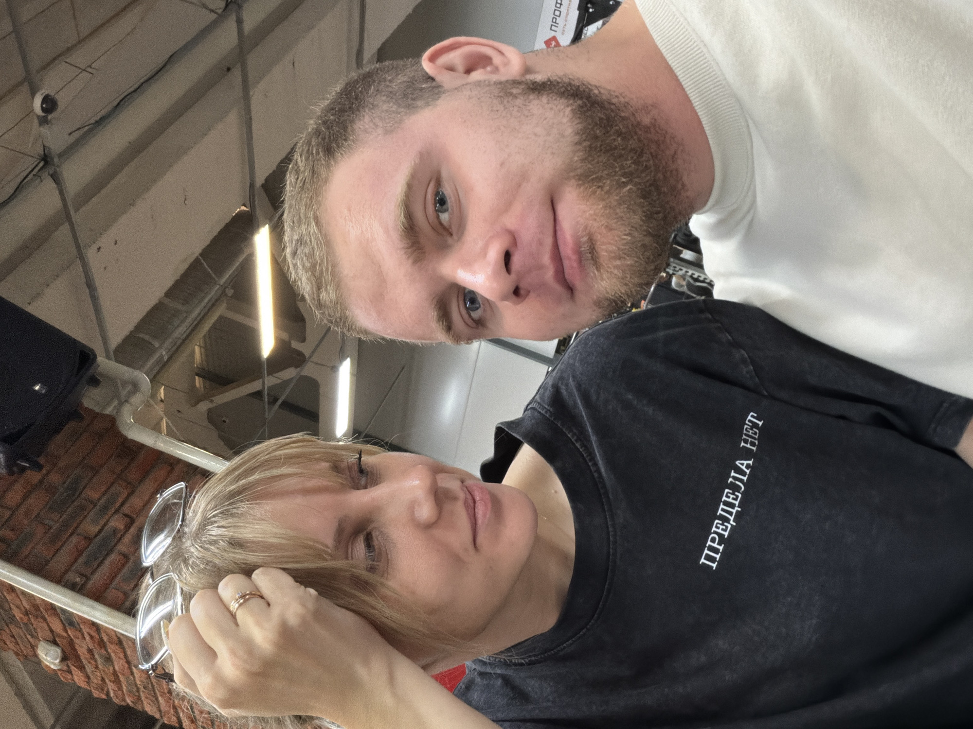 A selfie taken indoors shows two people. A light-skinned male with a beard and short hair is on top, wearing a white shirt. Below him is a light-skinned female with blonde hair, wearing a dark t-shirt with white text "ПРЕДЕЛІA НЕТ". The background shows a ceiling with lights, pipes, and brick elements. The location is potentially a commercial or industrial space in Chaikovsky, Russia.