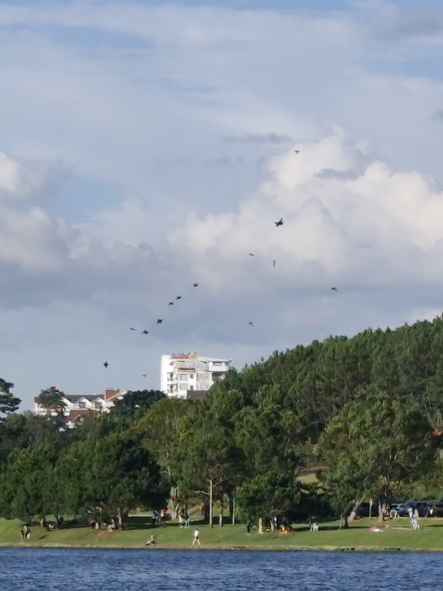The image depicts a panoramic view of Xuân Hương Lake in Da Lat, Vietnam. The foreground is dominated by the calm blue waters of the lake. The middle ground features a lush, green expanse of trees and grass lining the lake's edge. A few people are scattered across the grassy area, engaged in various leisurely activities, such as sitting, walking, and sunbathing. In the background, a large, multi-story white building with a distinctive architectural style stands out amidst the dense, green forest that covers the hillsides. The sky above is a mix of blue and white clouds, with several small objects, possibly kites or model airplanes, visible as dark specks against the sky. The overall scene conveys a sense of a peaceful, recreational day by the lake.