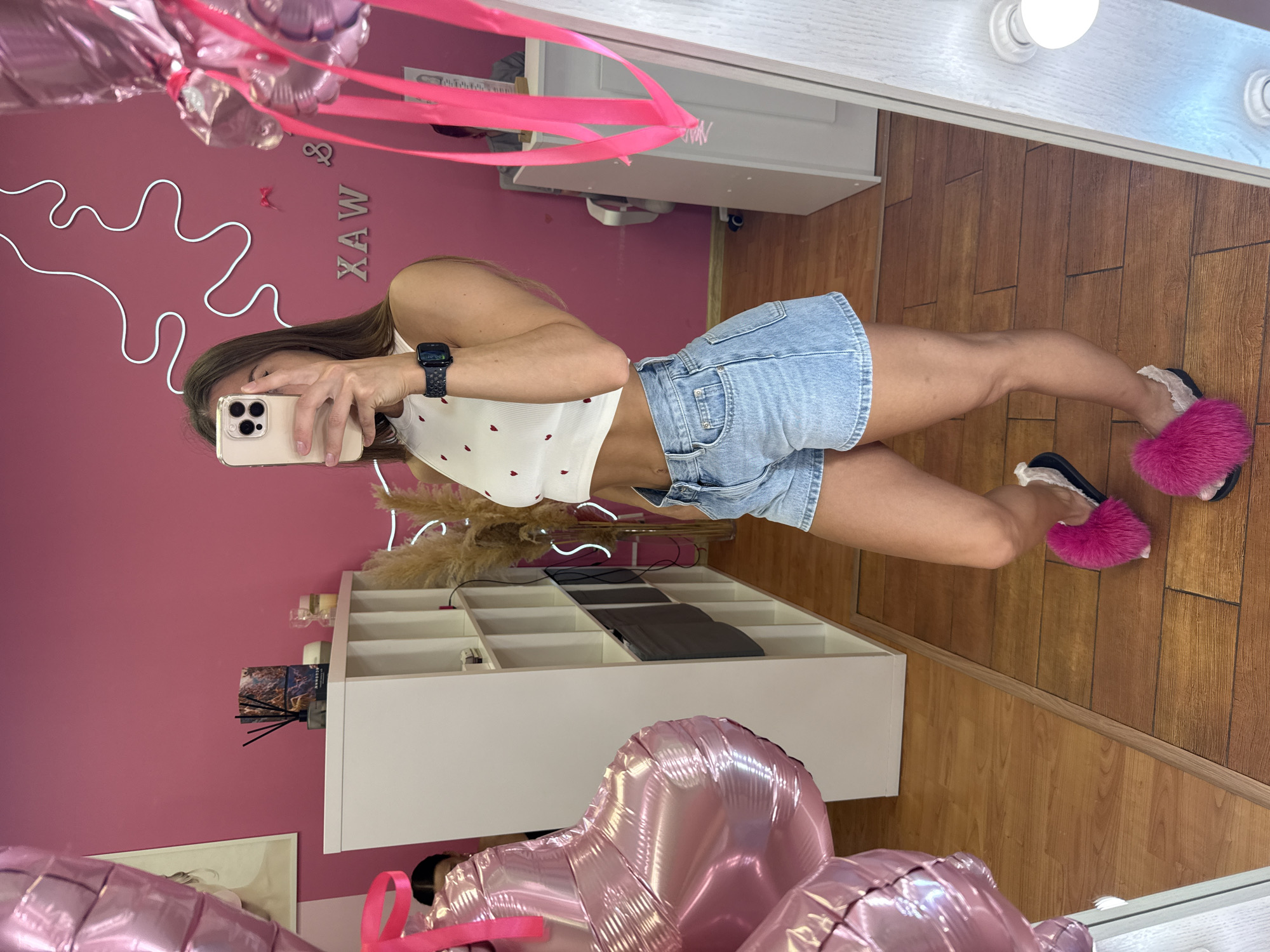 A woman is taking a mirror selfie in a room with pink walls and wooden floors. She is wearing a white crop top with small red hearts, denim shorts, and fluffy pink slippers. There are pink heart-shaped balloons to her left and a white shelving unit behind her. A sign with the letters "XAW" is on the wall. The room appears to be a salon, possibly for waxing services. The overall impression is one of a casual, personal photo being taken in a commercial space.