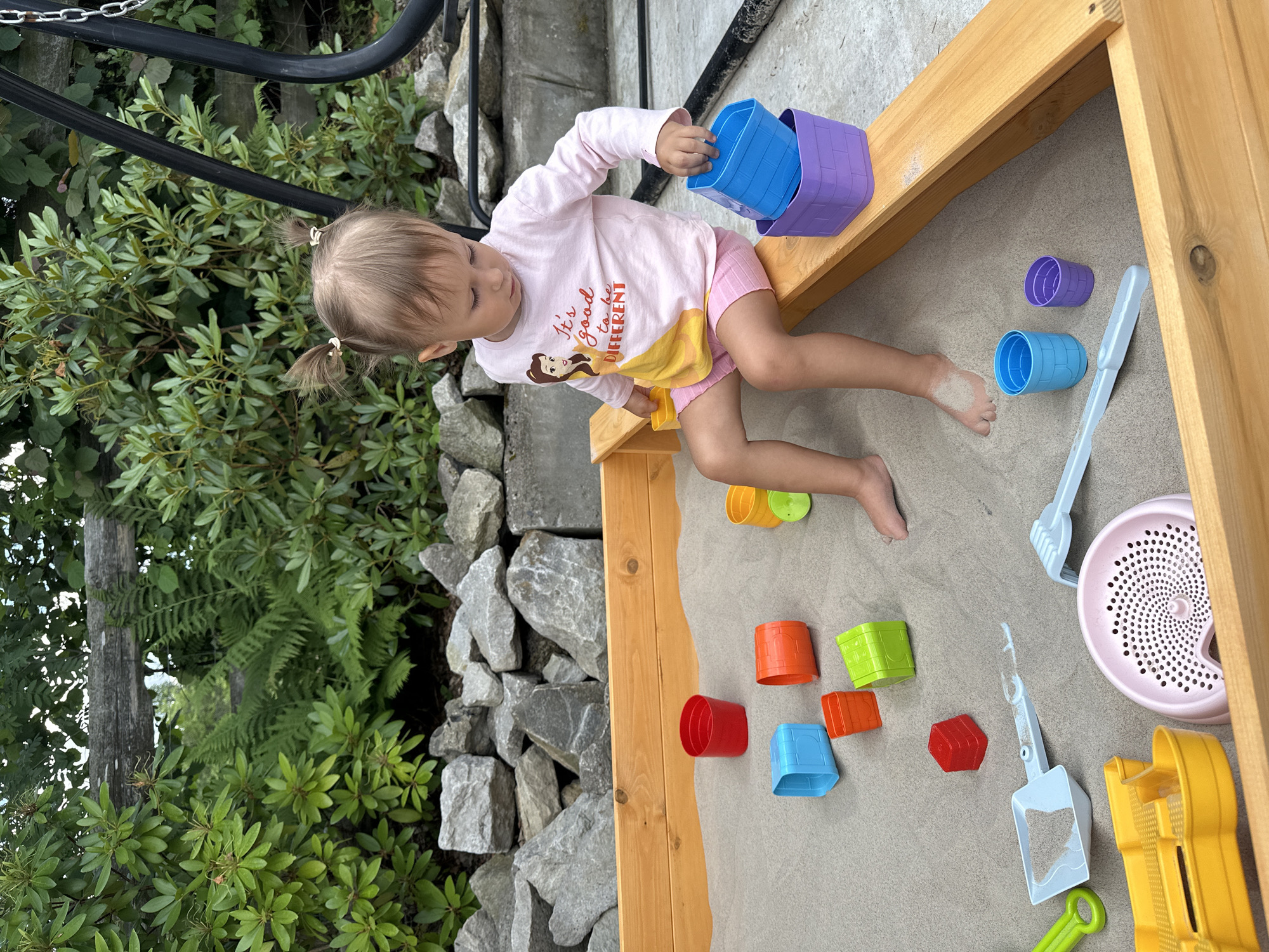 A young girl with blonde pigtails is sitting in a sandbox. She is wearing a pink long-sleeved shirt with a graphic of Belle from Beauty and the Beast and pink shorts. She is looking down at her hands as she stacks blue and purple plastic containers. The sandbox is made of light-colored wood and filled with sand. Scattered in the sand are various colorful plastic toys, including buckets, shovels, and molds. In the background, there is a stone wall and lush green foliage. The setting appears to be an outdoor play area, possibly a backyard. The image is shot from a slightly elevated angle.