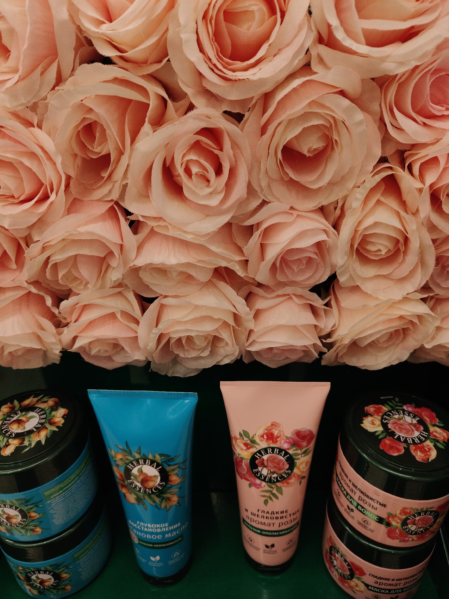 The image shows a collection of Herbal Essences hair care products arranged against a backdrop of numerous pale pink roses. The products consist of two types: tubs of hair mask and tubes of conditioner. The tubs are cylindrical with dark green lids and labels featuring the Herbal Essences logo and the name of the specific product line. Two of the tubs have a design with roses, while the other two have a design with argan nuts. The conditioner tubes are turquoise and pale pink, corresponding to the argan and rose product lines, respectively. The roses form a dense, artificial-looking wall above and partially behind the products, creating a soft and romantic visual effect. The setting implies a retail or promotional context, possibly within a beauty store or during a product launch.