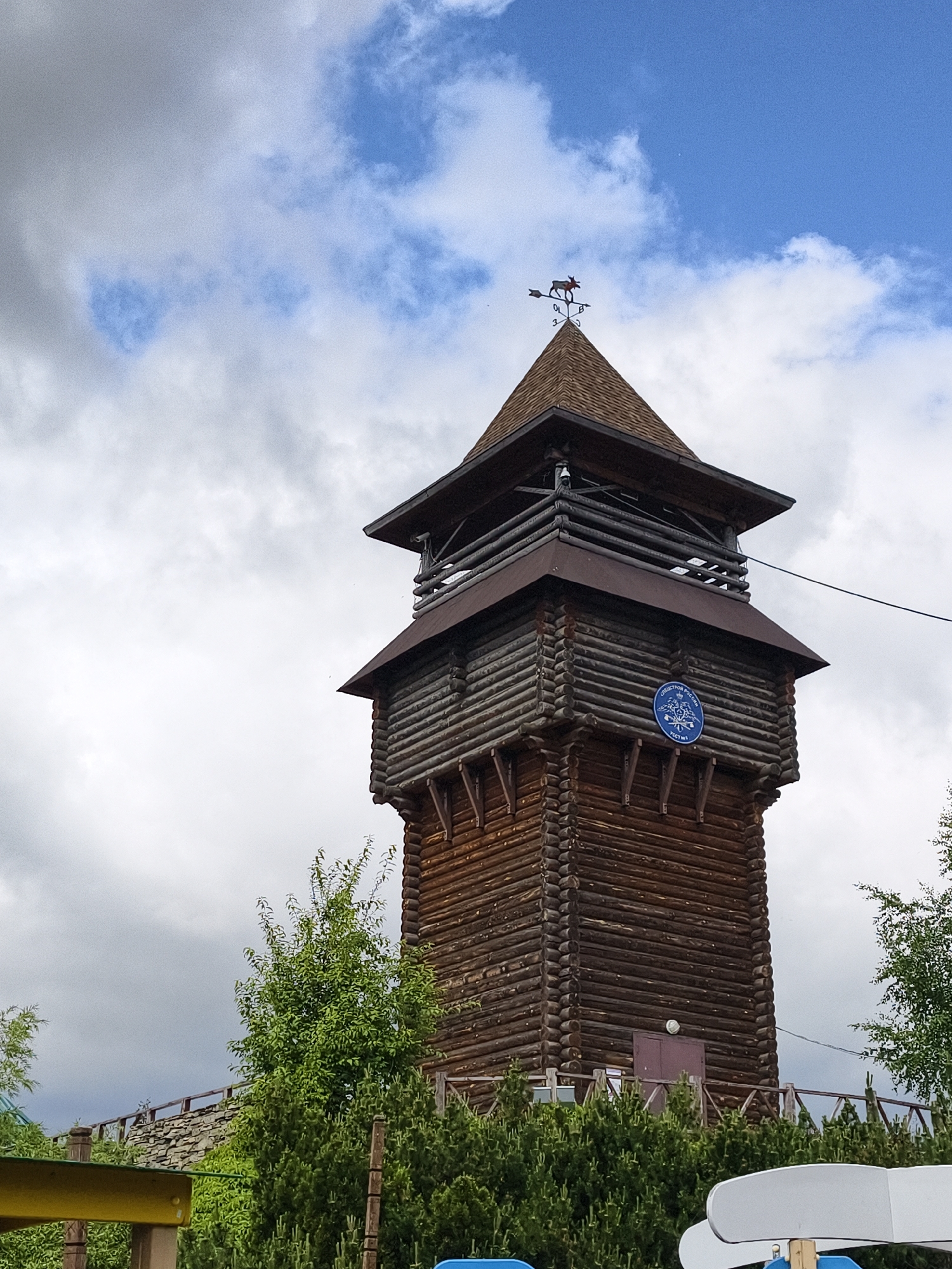 A tall, multi-level wooden tower, constructed from dark-stained logs, stands prominently under a partly cloudy sky with patches of bright blue. The tower features a wide, shingled roof with a smaller, second-tier roof below it, creating an observation or balcony area with log railings.
At the very top of the tower's spire, a weather vane with an animal figure, resembling a moose or elk, points to the left. Below the animal, directional markers are visible, though partially obscured. A thin wire or cable runs diagonally across the mid-section of the image, attached to the tower.
Midway down the tower, on one of its log walls, a round, blue sign with white text and a central emblem is affixed. The text clearly reads "ОБЩЕСТВО РОССИИ" (Society of Russia), with a second, smaller line of text below it, which appears to be "УСТ 1 В". Further down, a brown door is visible on the tower's base.
The tower is surrounded by lush green trees and bushes, with parts of a wooden fence or railing also visible. To the left, behind the greenery, a stone wall structure is present. In the lower foreground, parts of a yellow and a white structure, possibly playground elements, are visible. The scene is brightly lit, indicating daytime. No people or specific activities are depicted. The setting appears to be an outdoor recreational or historical park area in Izhevsk, Russia, with the tower's architecture and the visible Russian text serving as location cues.