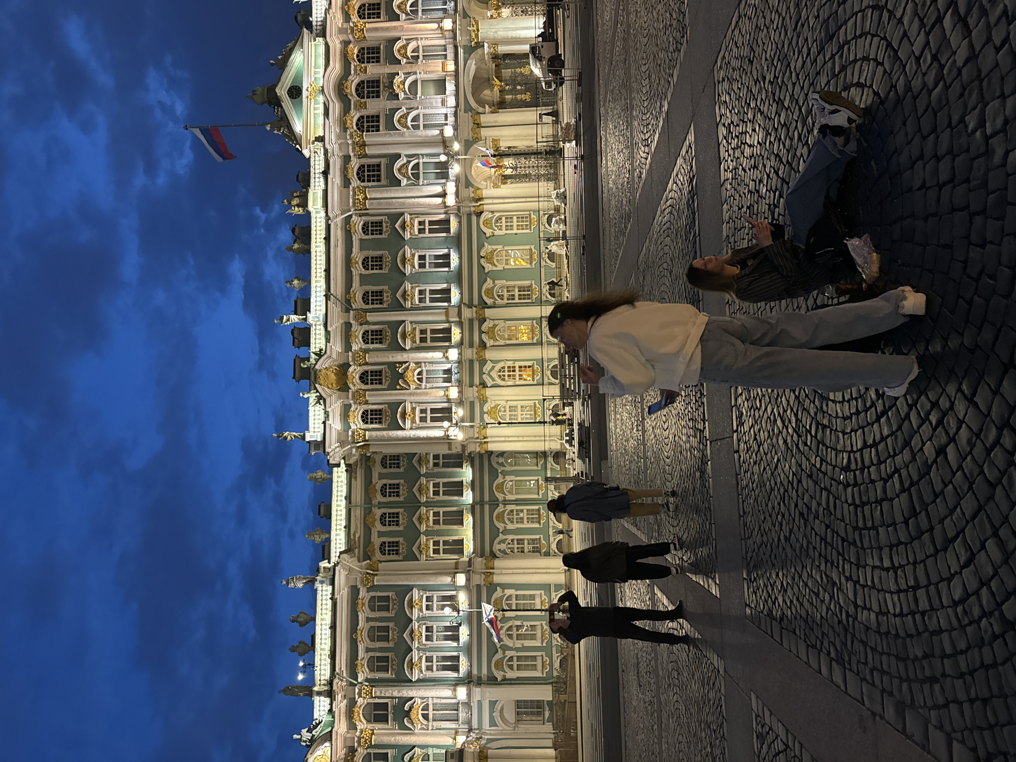 The image shows the Winter Palace, a historic building in Saint Petersburg, Russia, at twilight. The facade of the palace is illuminated, with its green and gold accents prominent against the darkening blue sky. Statues are visible along the roofline, and the Russian flag flies from a flagpole on top of the building.
In the foreground, several people are in the Palace Square, which is paved with cobblestones. Three individuals stand in the middle ground, one holding a phone to his face as if taking a photo. In the right foreground, two young women are seated on the ground. One is looking down at her phone, while the other is standing and appears to be drinking from a bottle or can. The overall atmosphere is one of evening tourism and observation of the grand architecture.