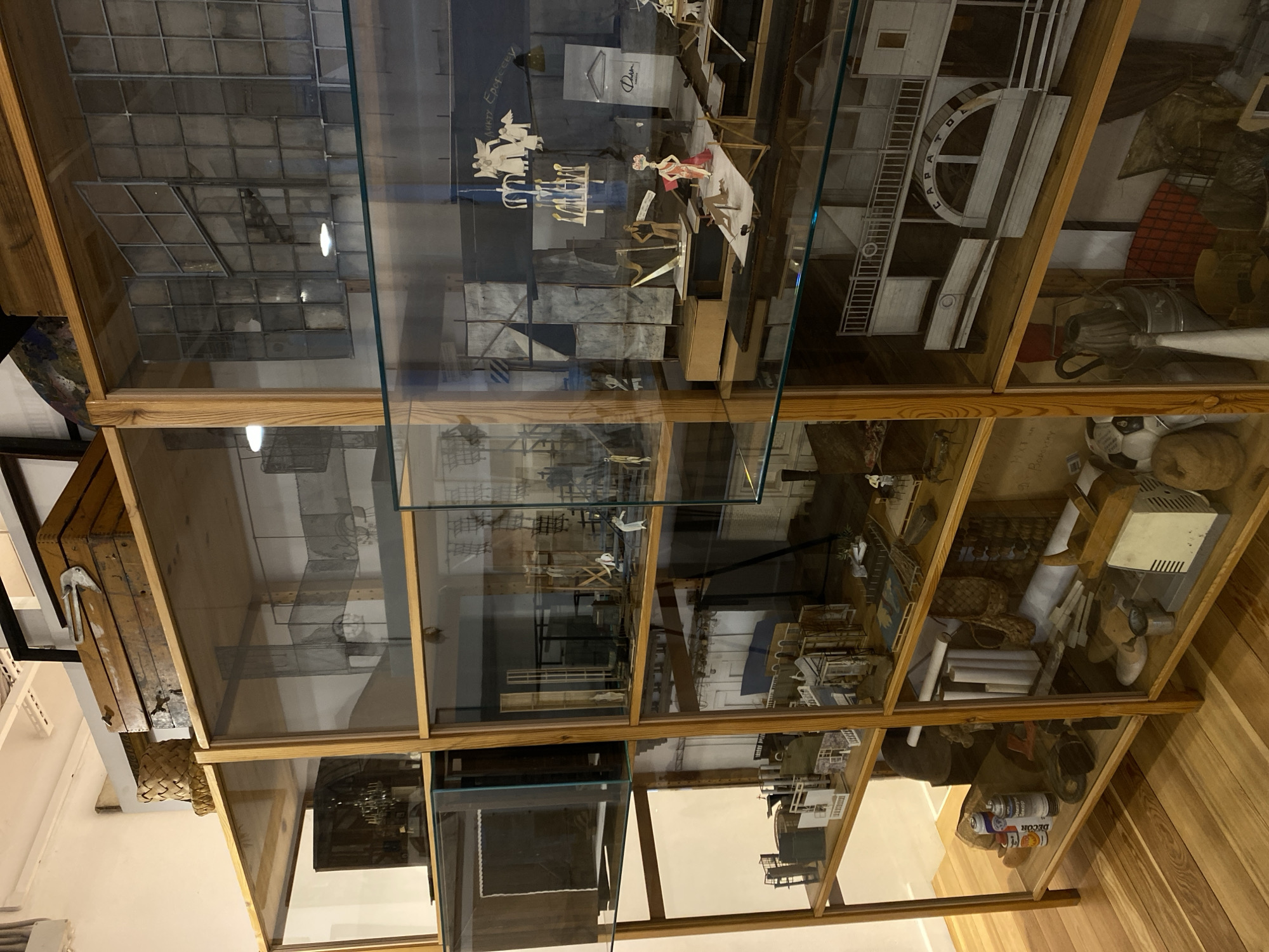 This media file captures a multi-compartment wooden display case with glass panels, likely situated in a museum or exhibition hall in Moscow, Russia. The case is filled with a diverse collection of objects, models, and artistic constructions.
In the upper sections, several intricate paper or cardboard cut-out figures are arranged, depicting groups of people, some in poses suggestive of dancing or theatrical performance. Behind these figures, faint Cyrillic text "АННА ЕРЕМЕЕВА" and the word "Dora" or "Dorn" are visible on a dark background. Another prominent exhibit is a detailed model of a white paddleboat or ship, featuring the clearly legible name "CARPATOL" on its side, accompanied by ladders and railings. Adjacent to this, there are architectural models, including structures resembling industrial buildings or parts of facades.
The lower compartments of the display case house an eclectic mix of items. These include rolls of paper, tightly woven baskets, a white electronic device with a vent, and various metallic components that appear to be vintage tools or machinery parts. In one section, several spray cans are visible, with "DECOR" clearly legible on one. Further down, a football (soccer ball) and other assorted objects, including a rustic wooden cradle-like structure, are displayed.
The scene portrays a curated exhibit, showcasing a wide range of subjects from detailed architectural and industrial models to artistic paper cut-outs and everyday objects. The indoor setting is illuminated by artificial lighting, with reflections of the ceiling's grid pattern and light sources visible on the glass panels. The floor outside the case is made of light-colored wooden planks. No individuals are present in the image, allowing an unobstructed view of the displayed artifacts.