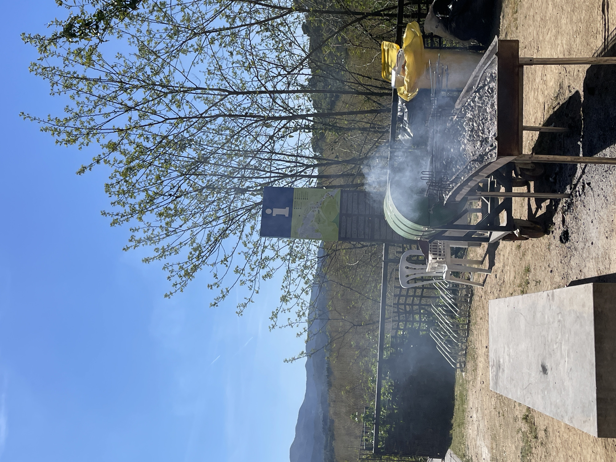 The image depicts a barbecue area in Hostalric, Spain. Visible is a smoking grill, with a container of ash or charcoal next to it. A yellow plastic bag is positioned near the grill. A sign with a blue "I" (information) is visible, along with a map. A white plastic chair sits nearby. The ground is dirt, and there's a concrete table. In the background, there are trees, a fence, and distant hills under a blue sky with sparse tree branches.